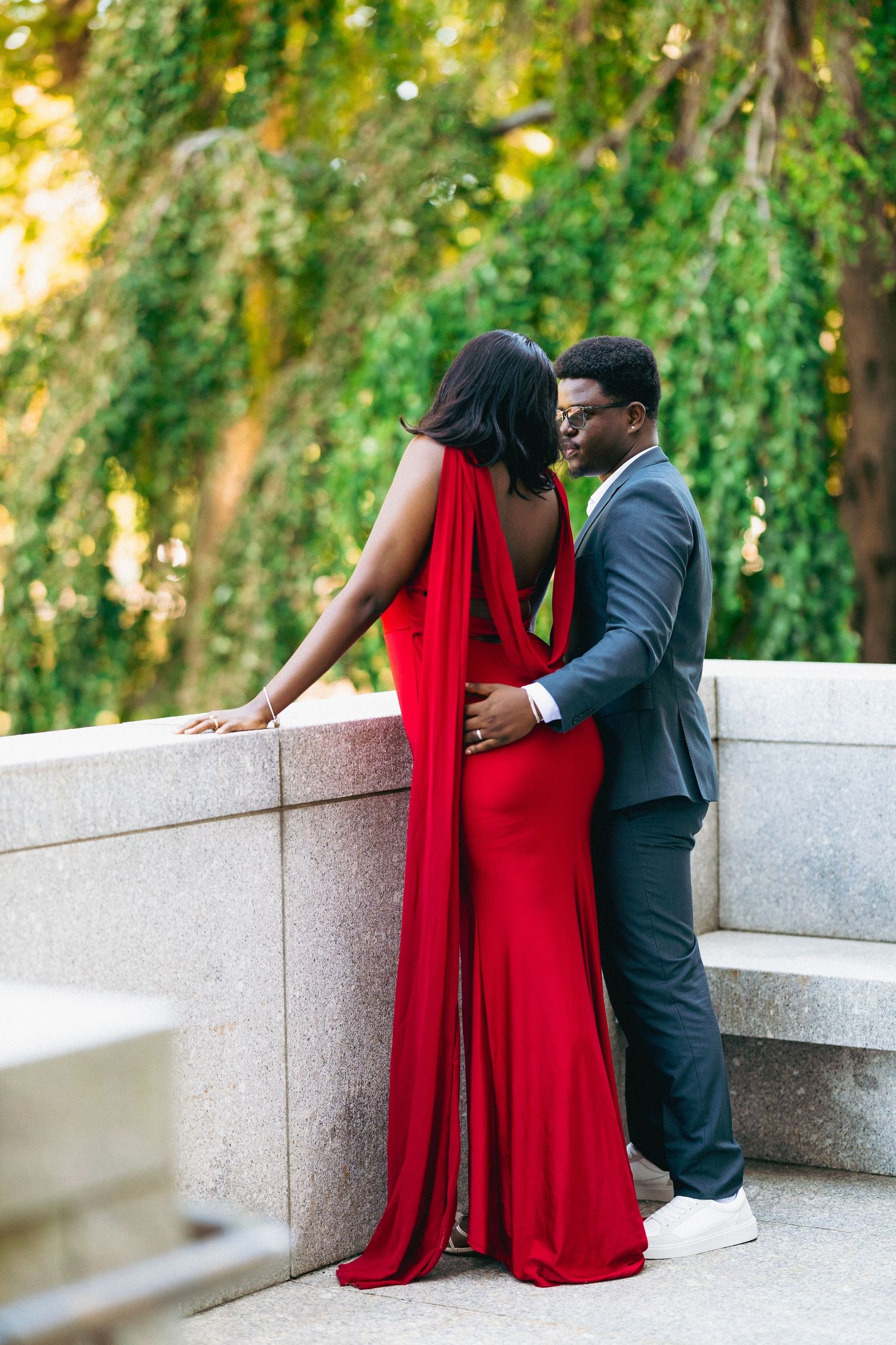 A couple in elegant attire, woman in red backless gown and man in gray suit, share a romantic moment outdoors.