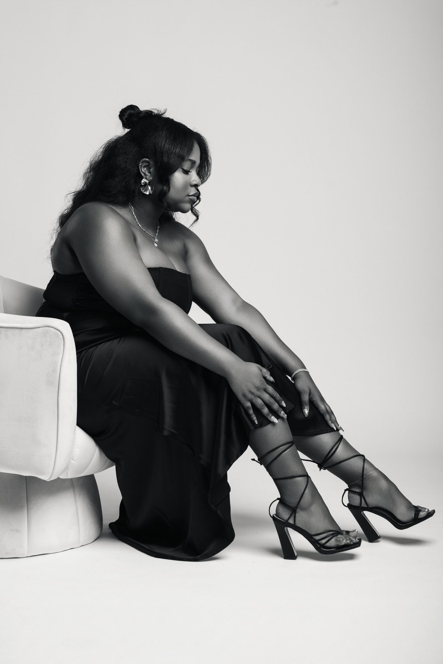 Black and white photo of a woman in a strapless black gown seated, wearing strappy heels.