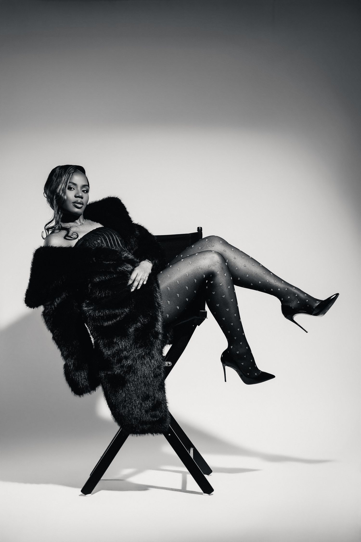 Black and white photo of a Black woman in a fur coat and heels posing in a chair dramatically.