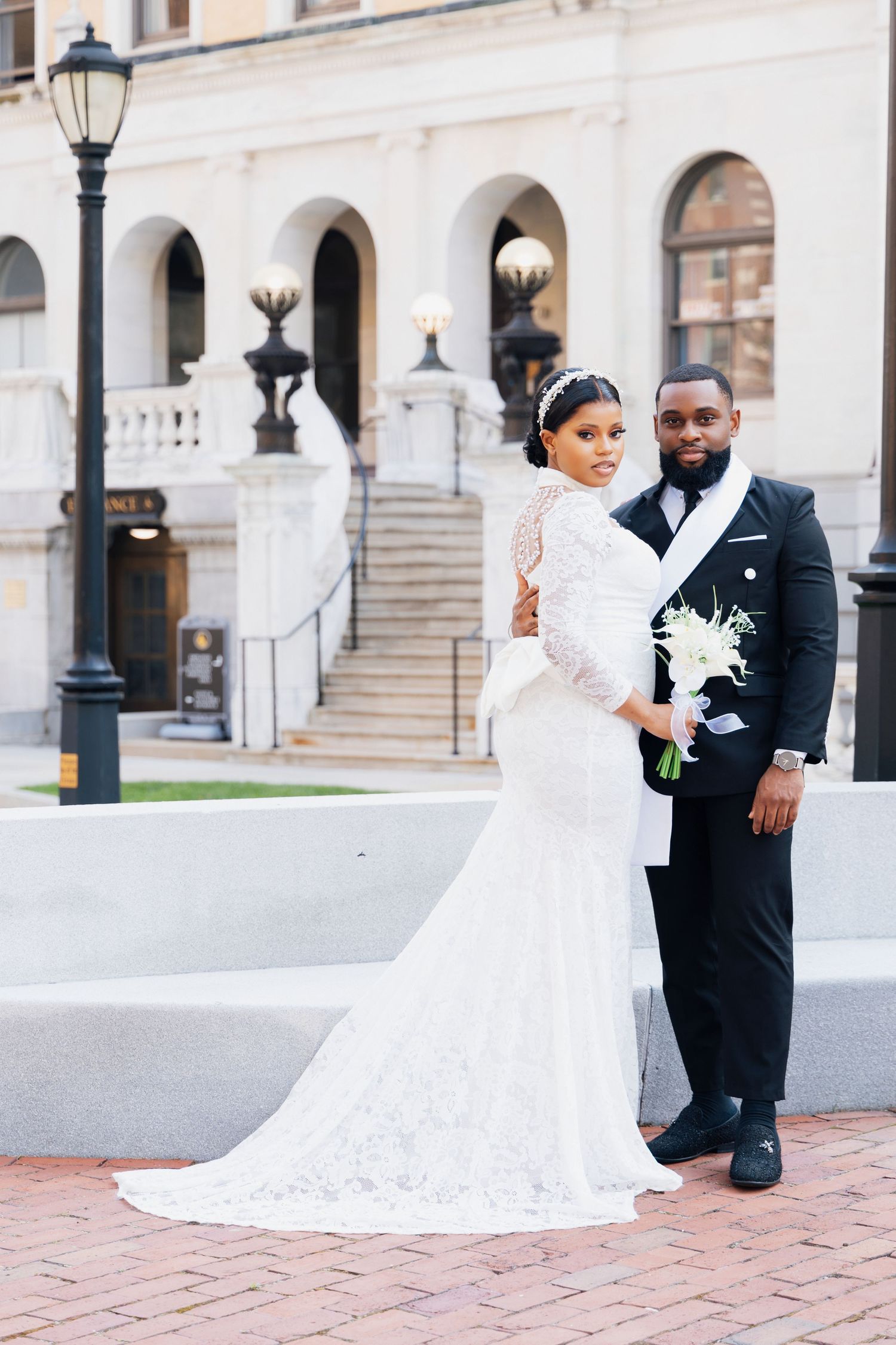 Elegant bride and groom posing in front of a grand building, bride in lace gown with train, groom in black tuxedo.