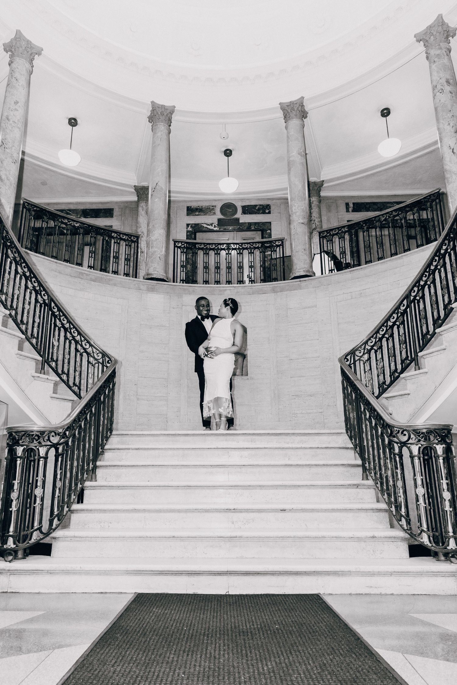 Black and white wide shot of bride and groom standing together on grand marble double staircase.