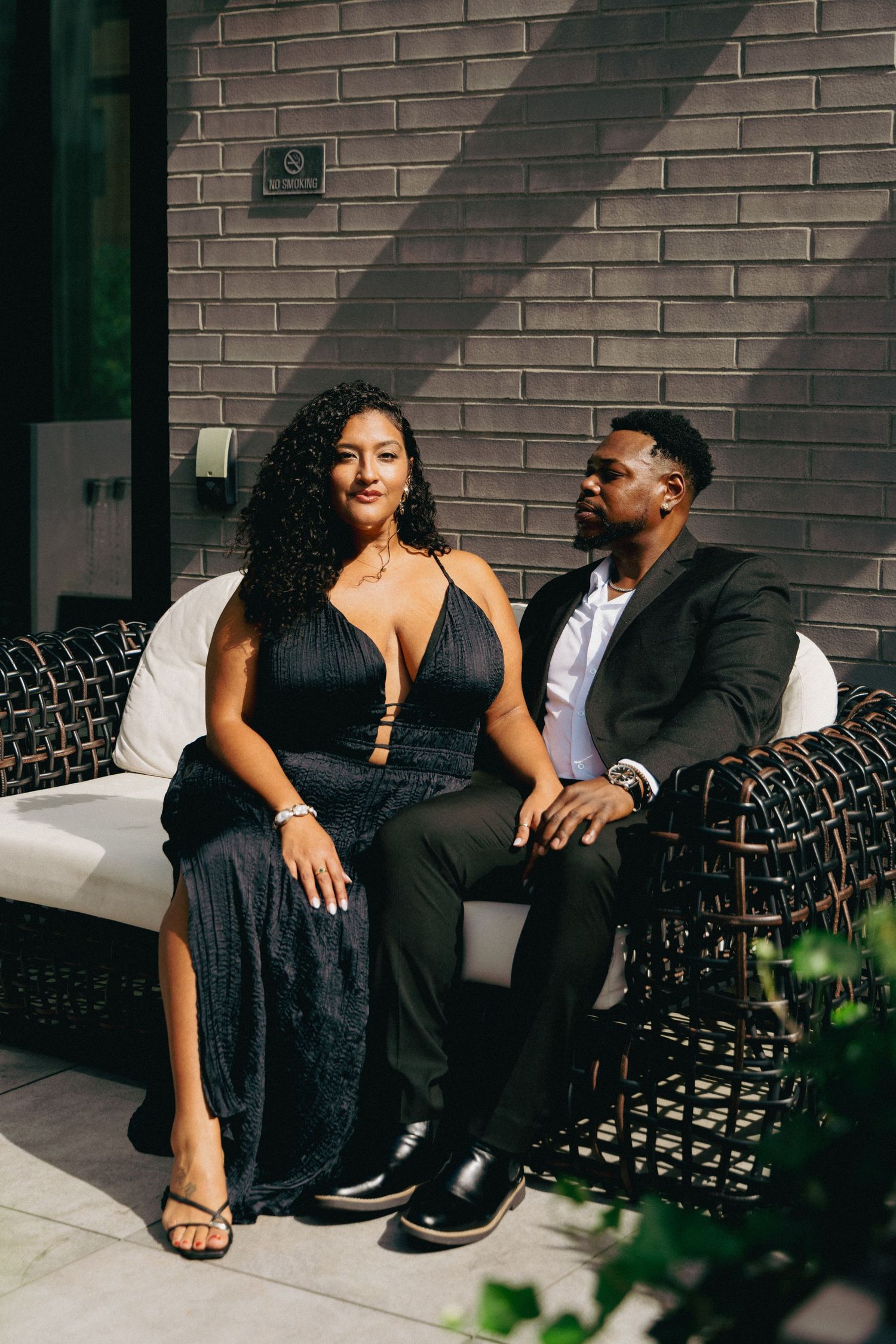 Elegant couple in black formal attire sitting together on wicker outdoor furniture against a brick wall.