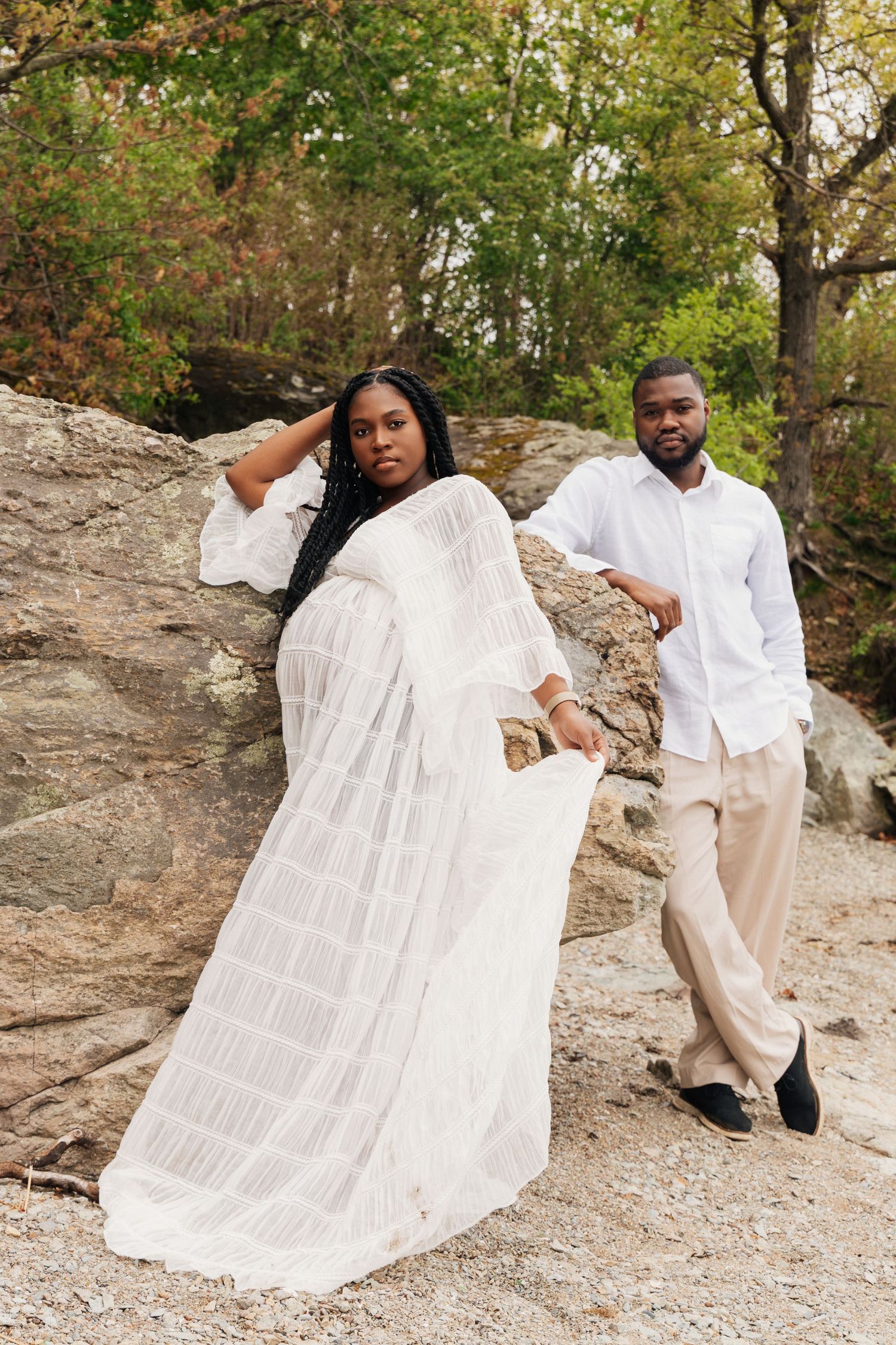 A Black couple poses against rocky terrain, woman in flowing white dress, man in white shirt and beige pants, surrounded by trees.