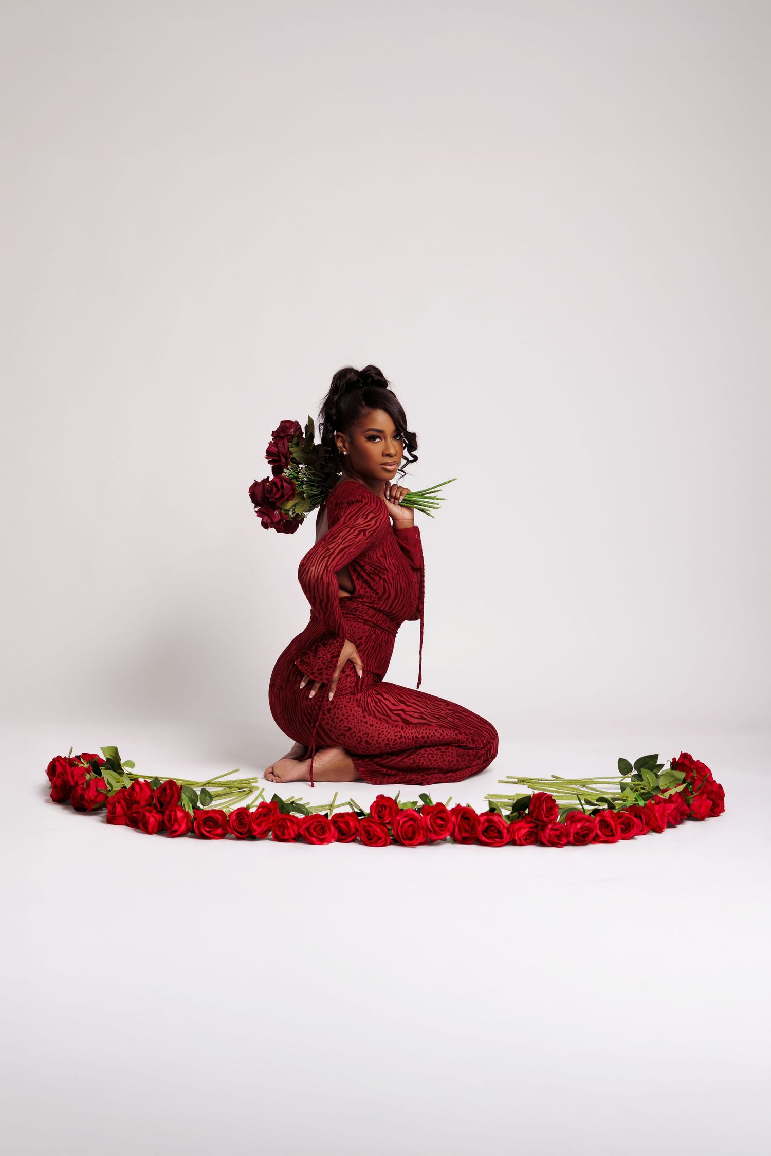 Woman in red knit dress sits gracefully surrounded by a semicircle of red roses, holding a bouquet.