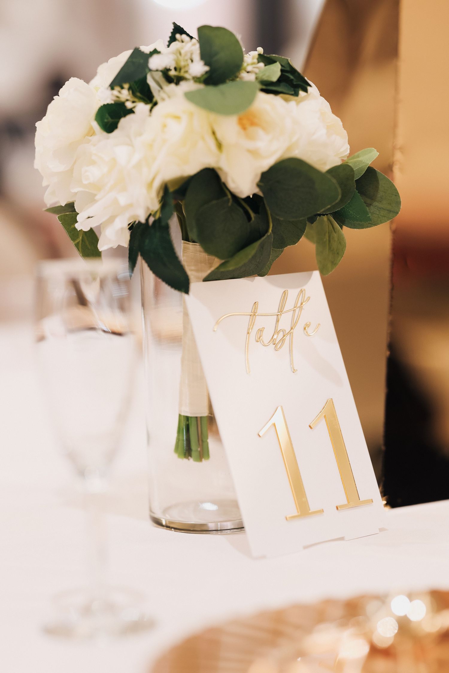 Elegant wedding table centerpiece with white roses and greenery in glass vase beside gold table number 11 card.