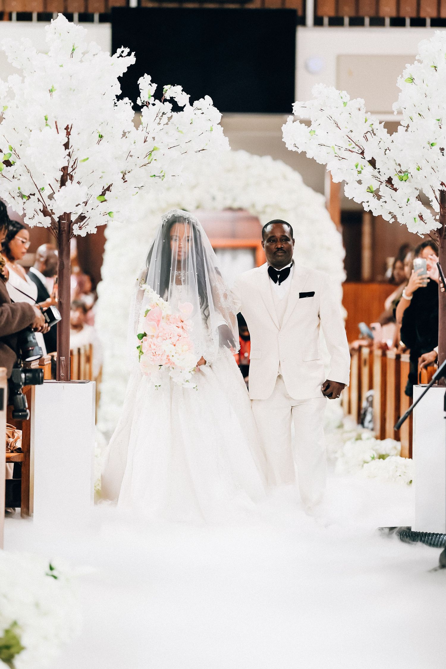 Bride in white gown and veil walks down the aisle with her escort at a church wedding ceremony.