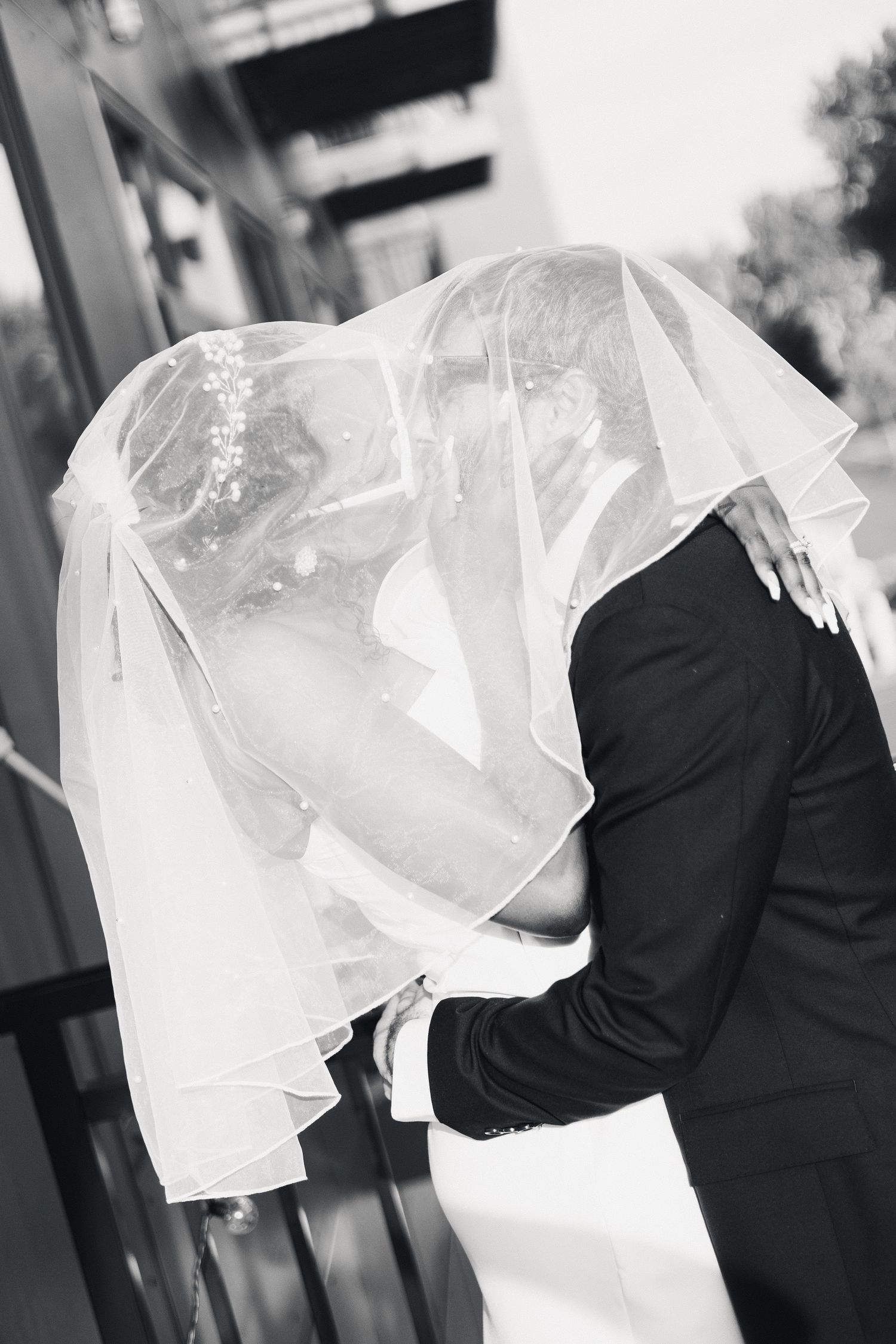 Black and white photo of newlyweds sharing a kiss hidden beneath the bride's flowing wedding veil outdoors.