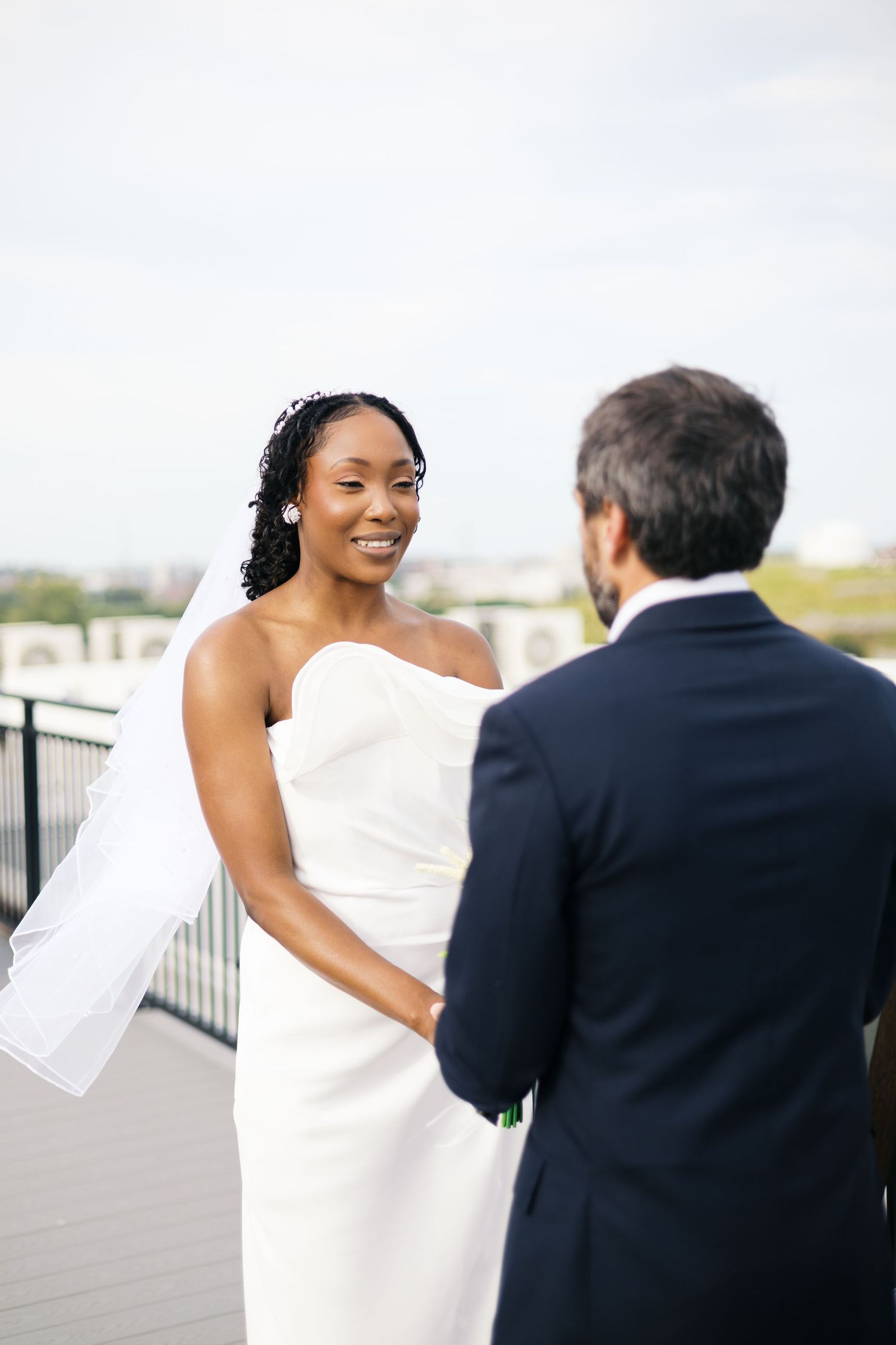 Smiling bride in white strapless gown and veil holds hands with groom in navy suit during outdoor rooftop wedding ceremony.