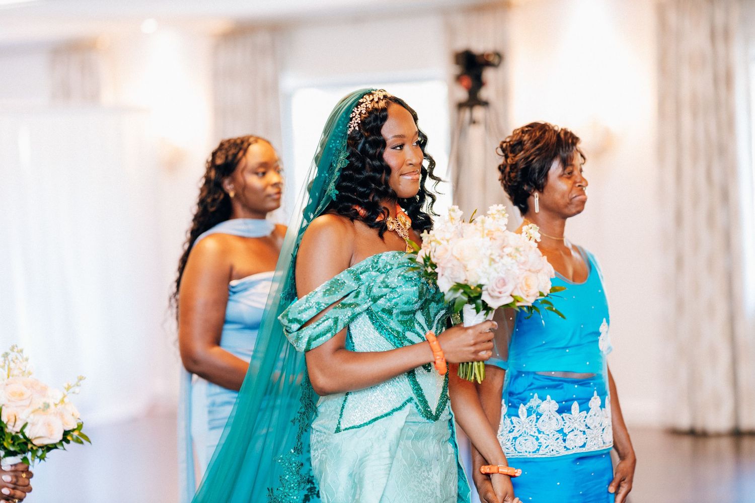 Bride in teal gown holds white bouquet flanked by bridesmaids in blue dresses at elegant wedding ceremony.