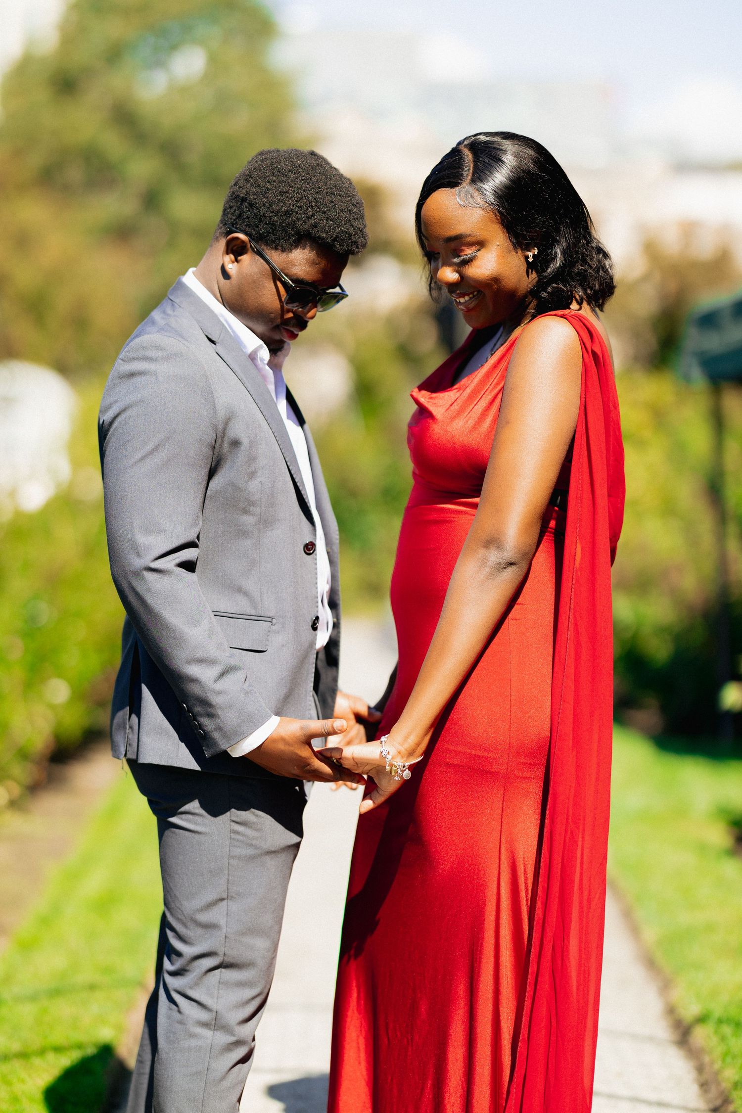 Elegant couple holding hands in a garden, woman in flowing red dress and man in gray suit smiling together.