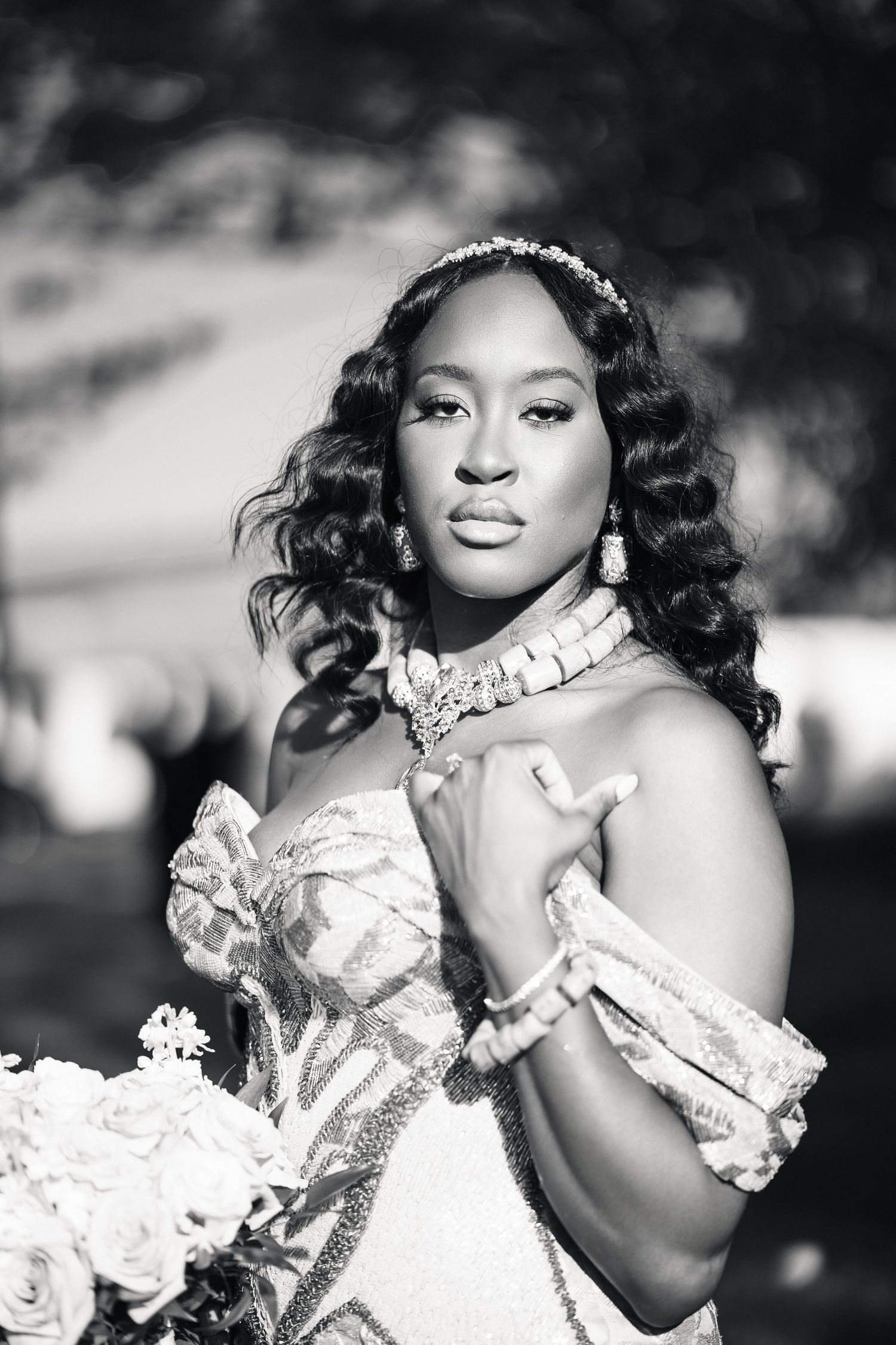 Black and white bridal portrait of an elegant bride wearing a jeweled headpiece, statement necklace, and embellished gown holding flowers.