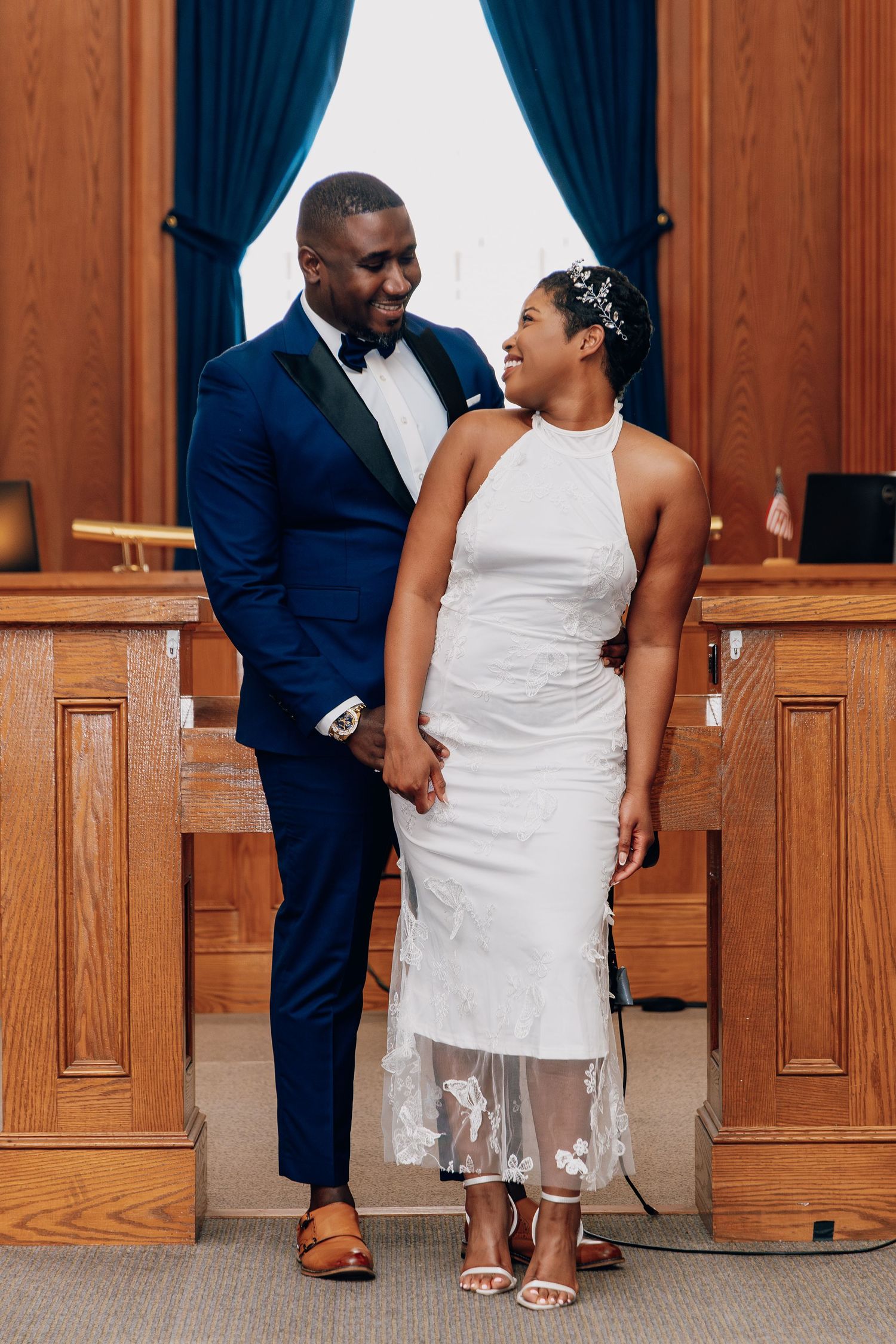 A joyful Black couple in wedding attire hold hands and gaze at each other in an elegant wood-paneled ceremony room.