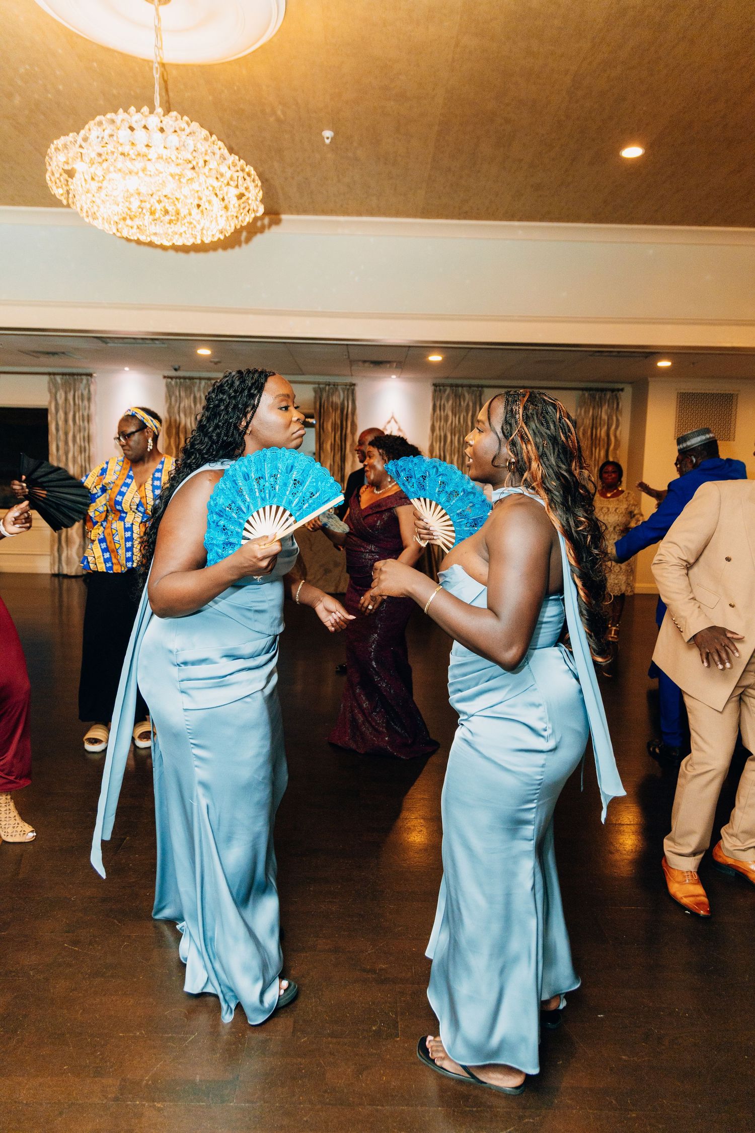 Two bridesmaids in matching light blue satin gowns dancing with decorative blue fans at a wedding reception.
