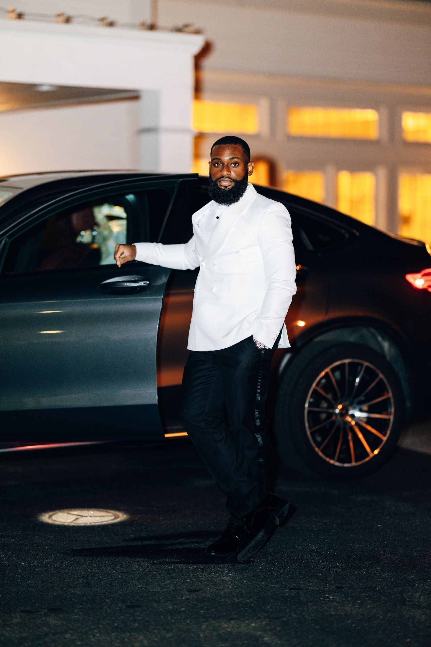 Stylish man in white shirt and black pants stands beside a dark luxury Mercedes car at night.