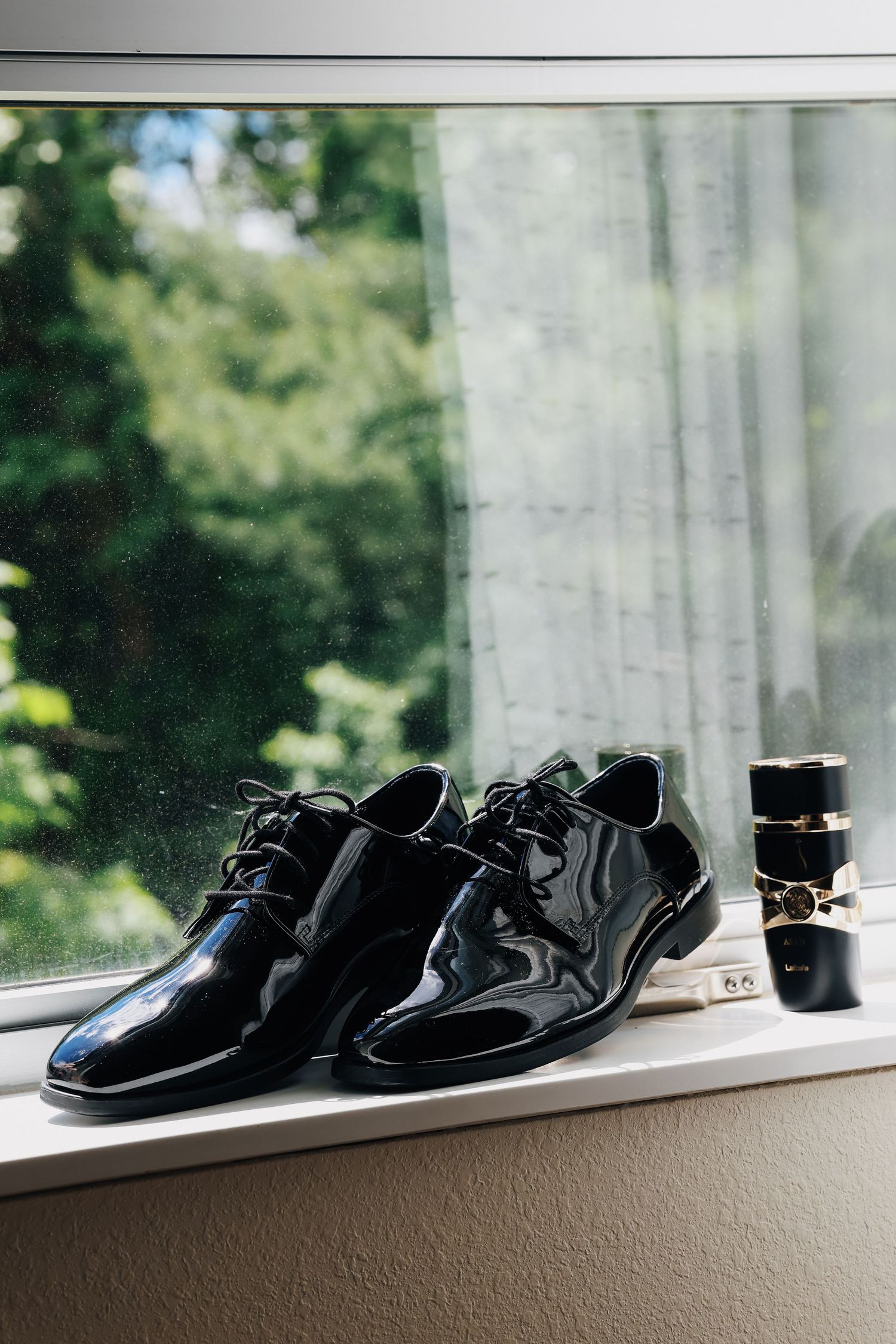 Black patent leather dress shoes and cologne on a windowsill with green trees in the background.