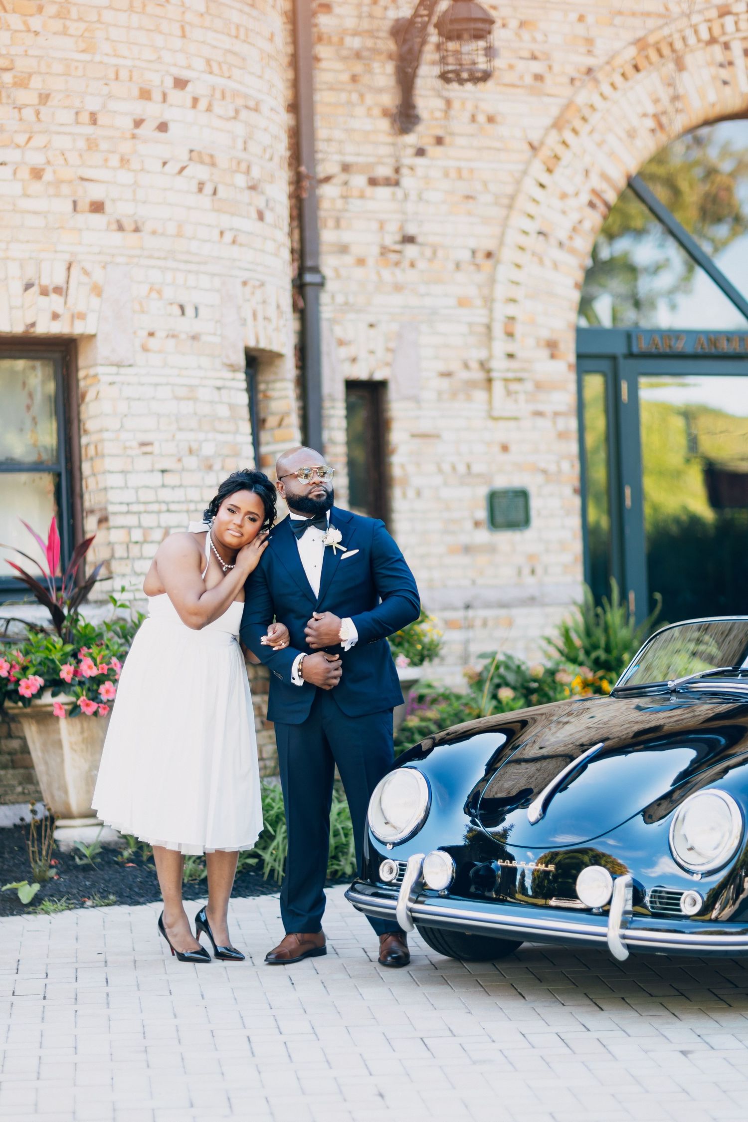 Newlywed couple standing together beside a classic black Porsche in front of a rustic brick venue.