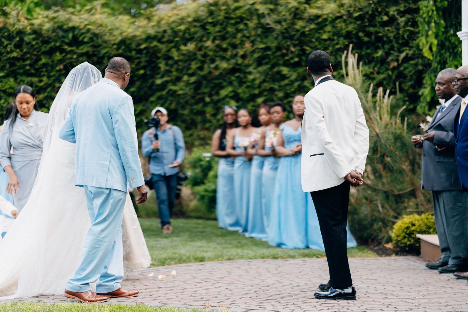 Outdoor wedding ceremony with bride in white gown, groom in light blue suit, bridesmaids in blue dresses.