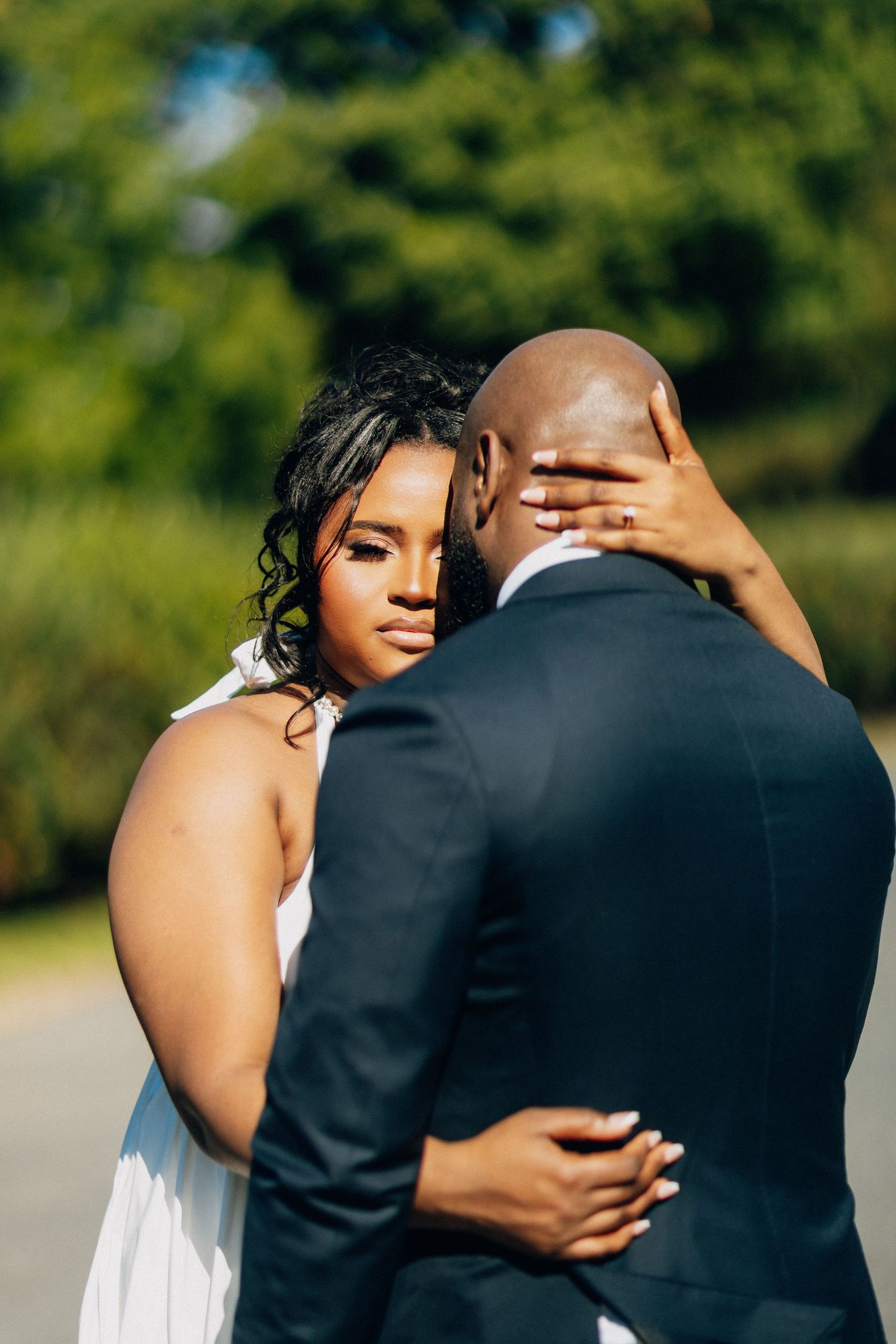 Bride in white dress embraces groom in navy suit outdoors, resting her hand on his head.