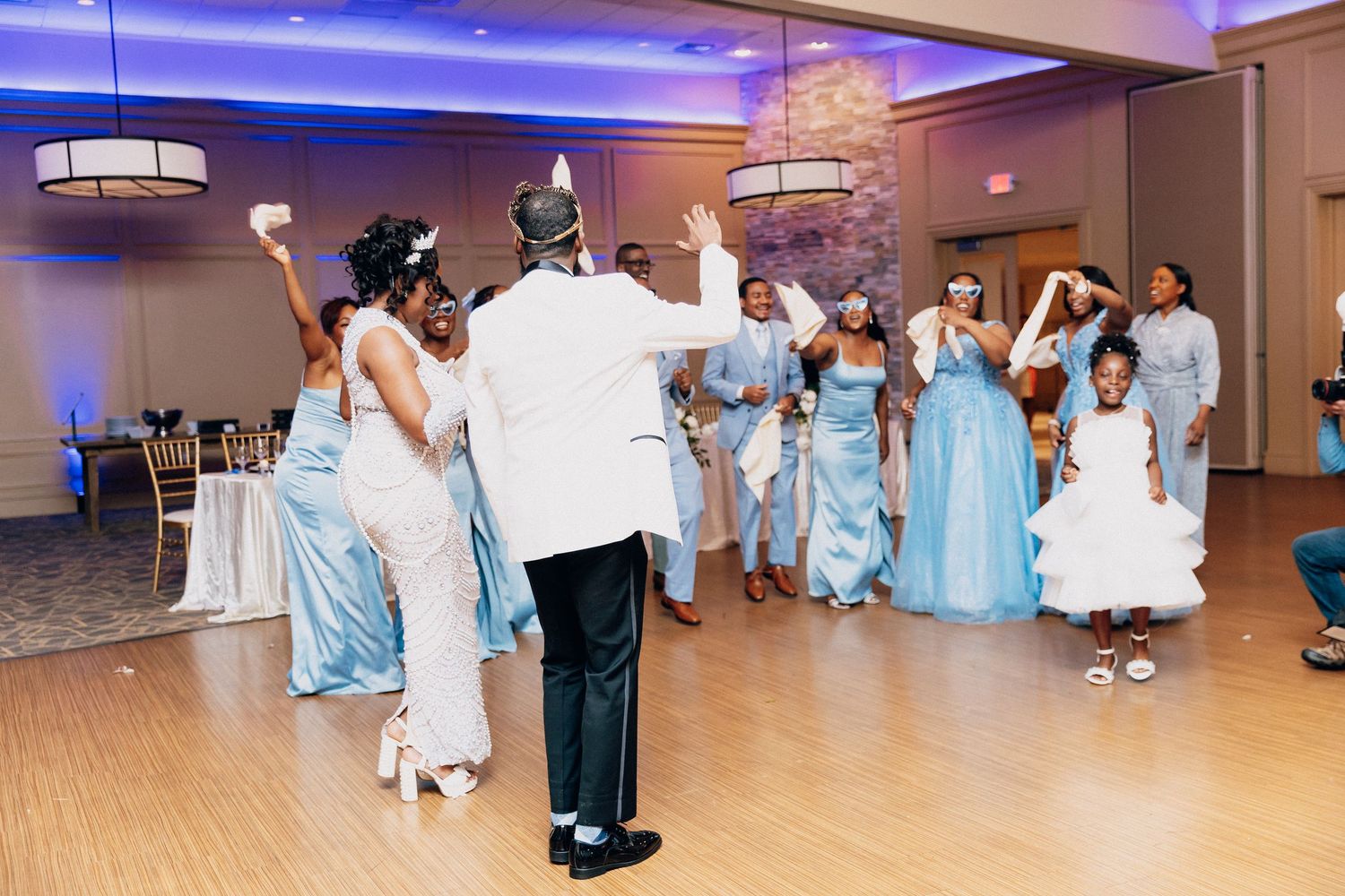 Wedding party in blue dresses dancing joyfully at elegant reception hall with purple ambient lighting.