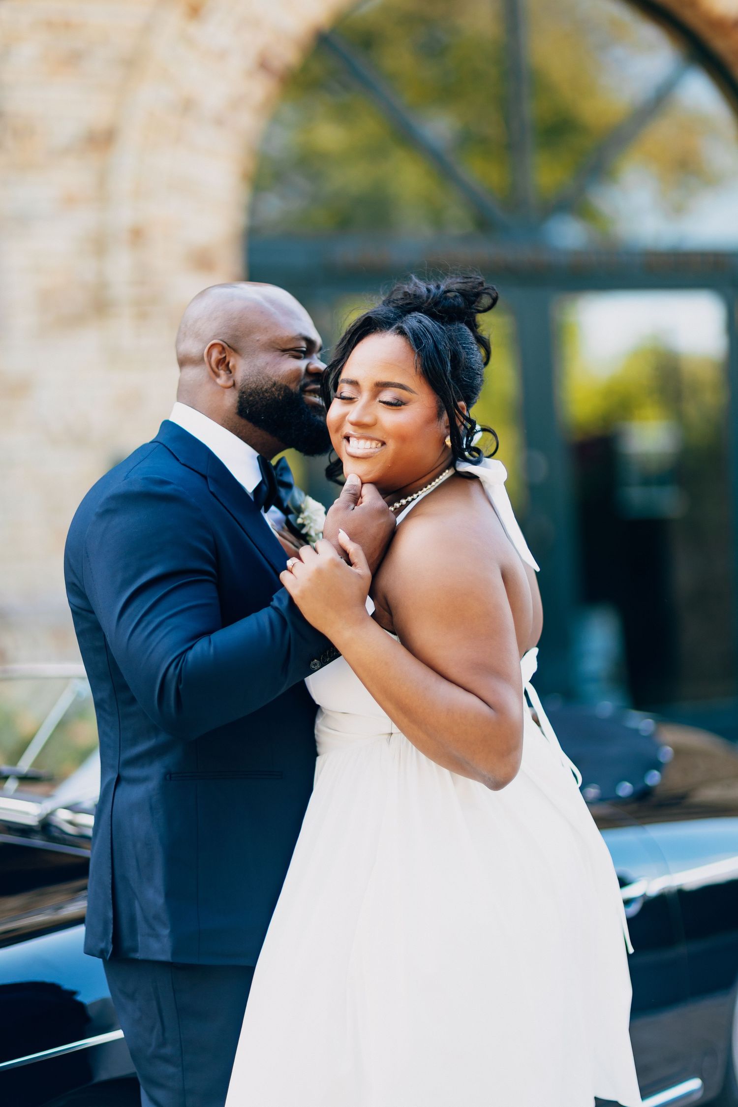Groom whispers to smiling bride near a vintage black car outside a stone wedding venue.