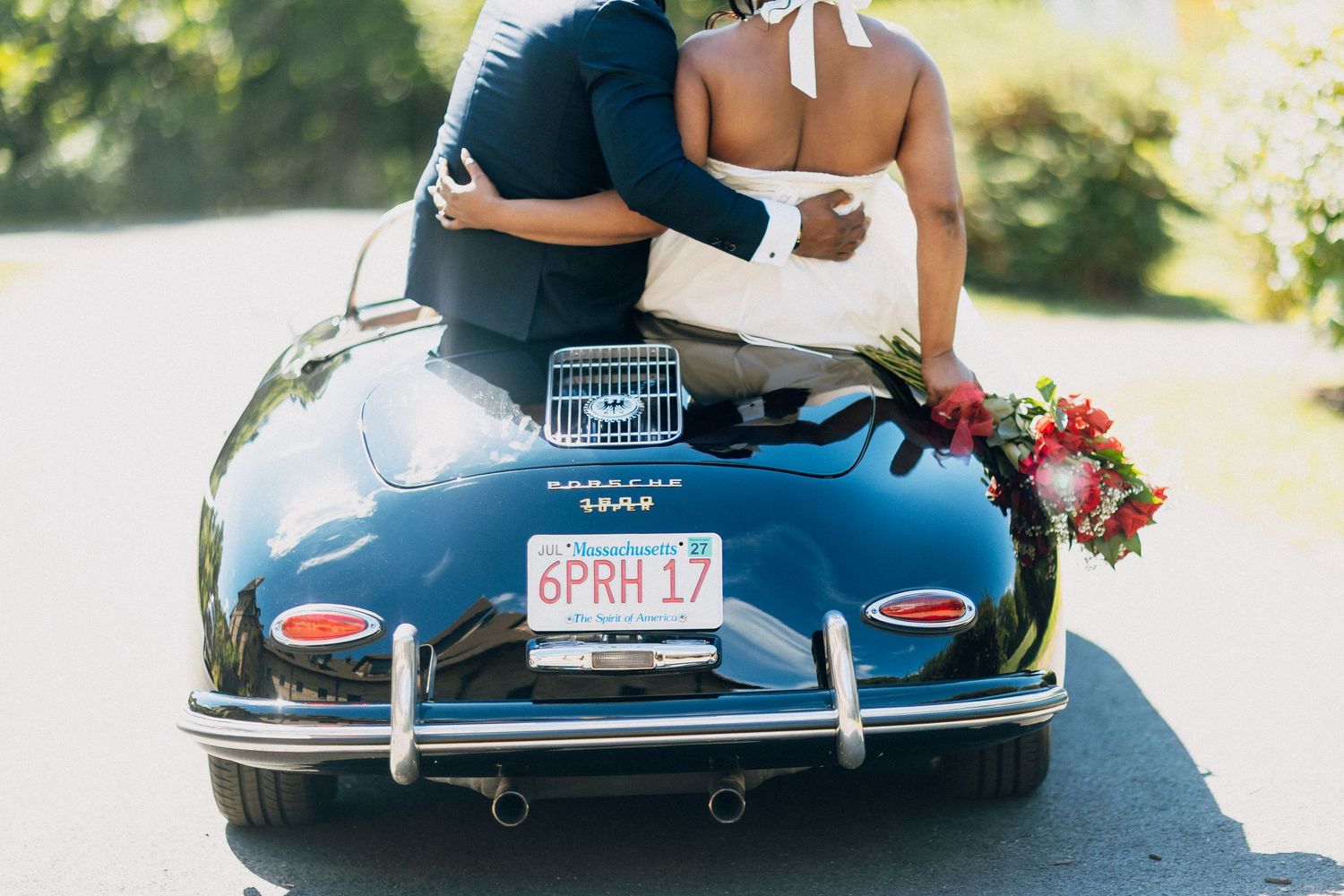 Newlyweds embracing on the trunk of a classic black Porsche 356 with a red floral bouquet.
