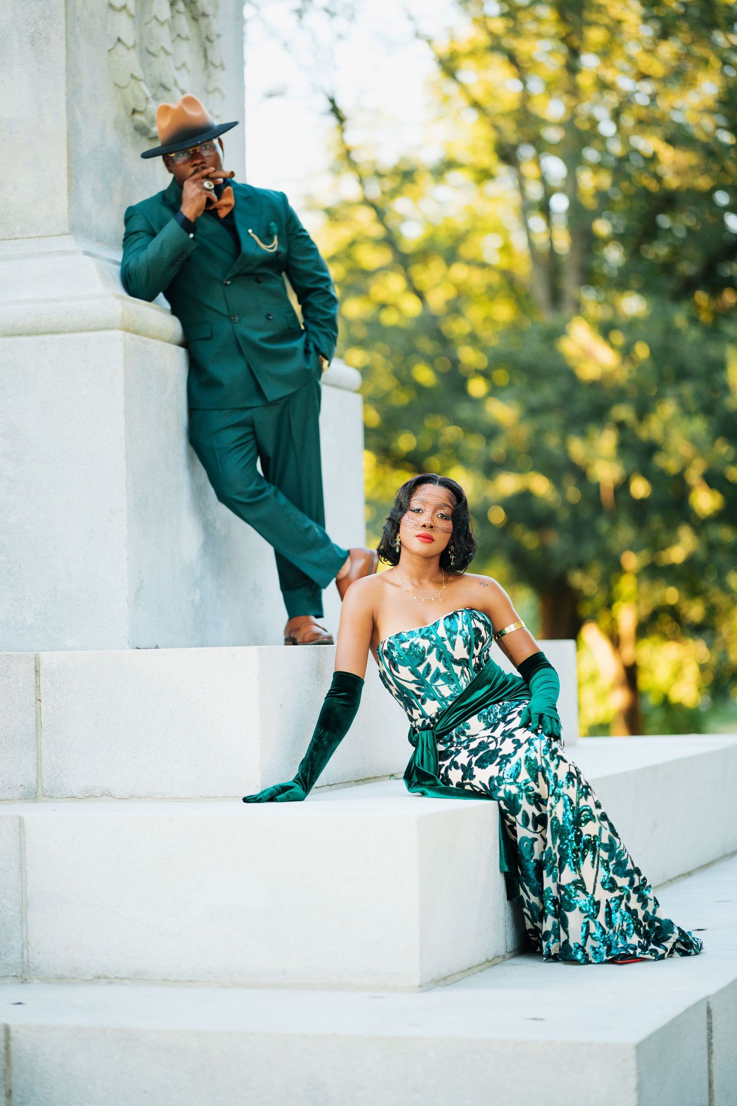 Sophisticated couple in emerald green attire posing on white marble steps surrounded by lush greenery.