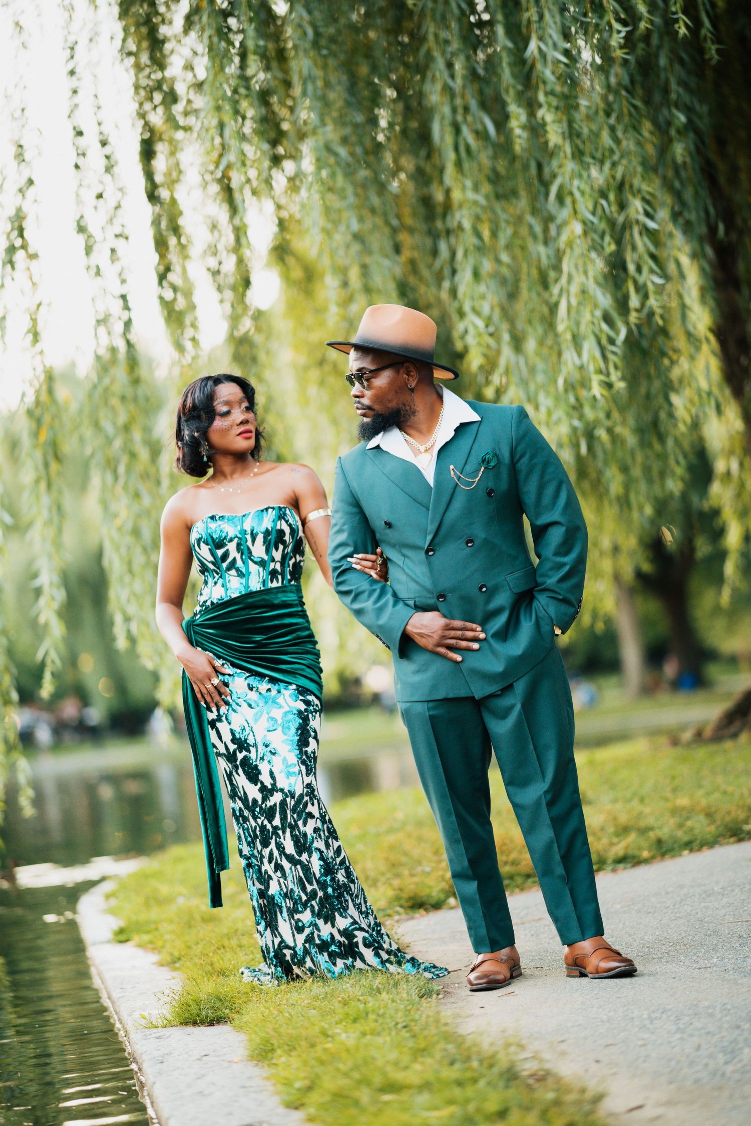 Fashionable couple in matching teal outfits walking along a park path near a pond under weeping willow trees.