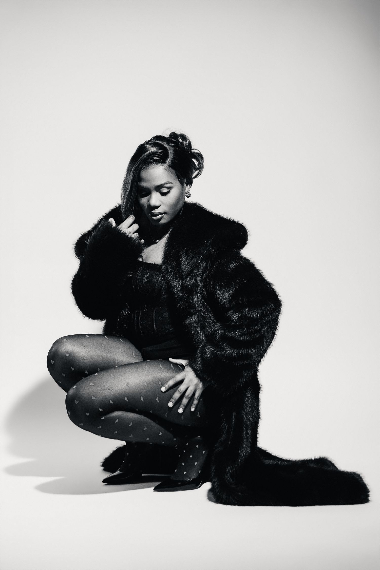 Black and white photo of a woman in a fur coat and patterned tights crouching gracefully in a studio setting.