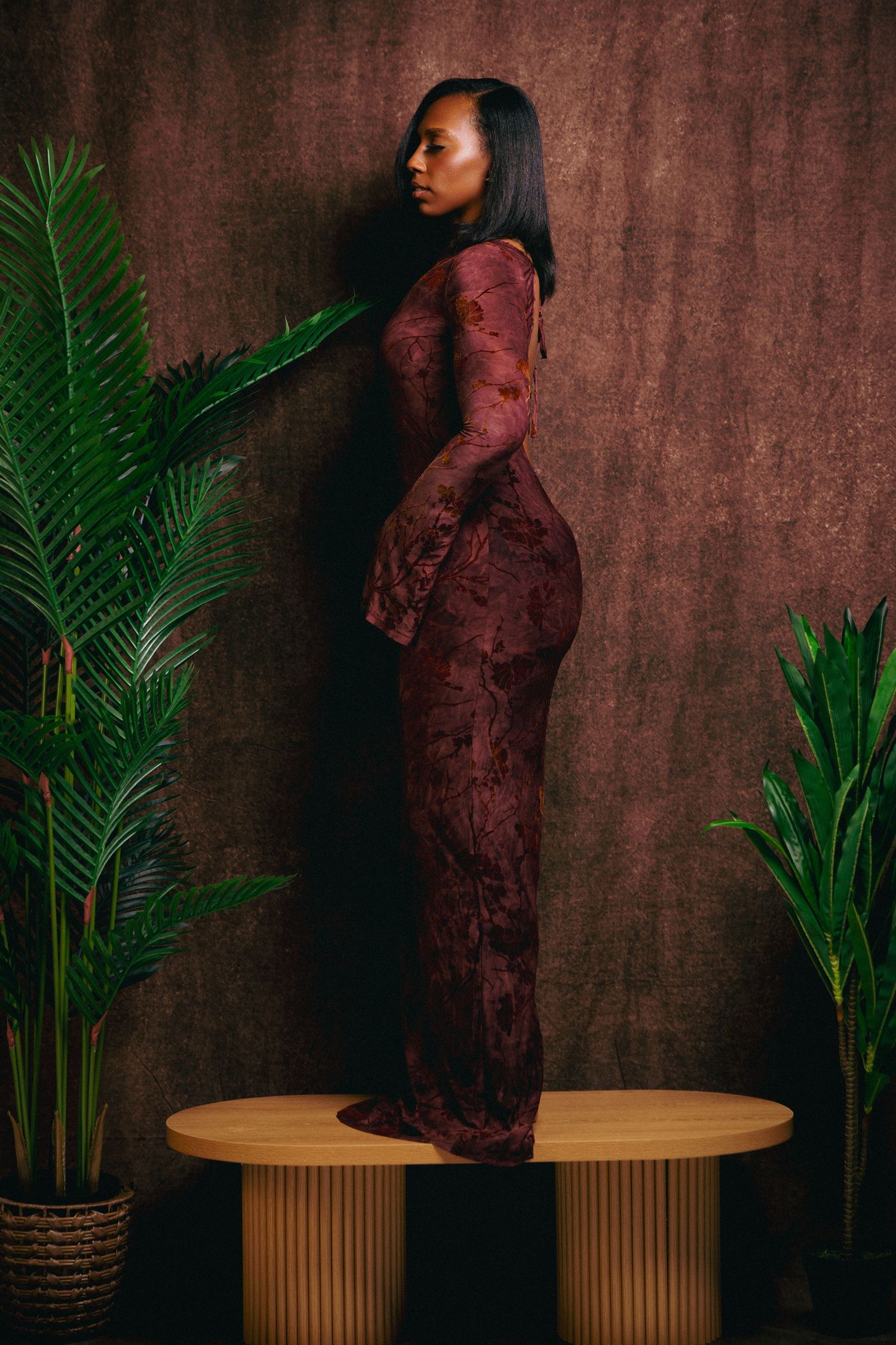 A Black woman in a burgundy lace maxi dress stands on a wooden pedestal beside tropical plants against a dark backdrop.