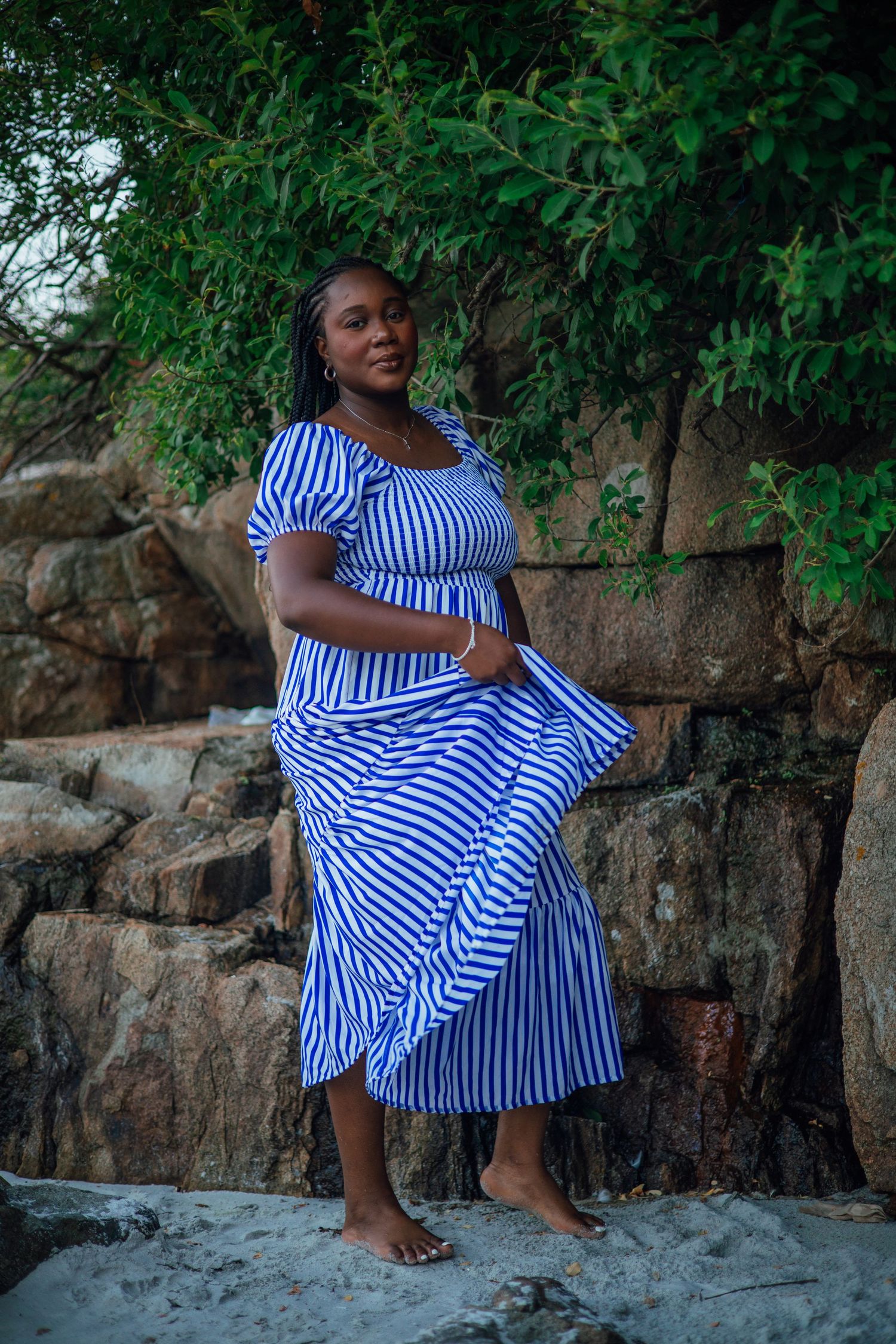 A woman in a blue and white striped maxi dress poses barefoot on sandy rocks with lush green trees behind her.