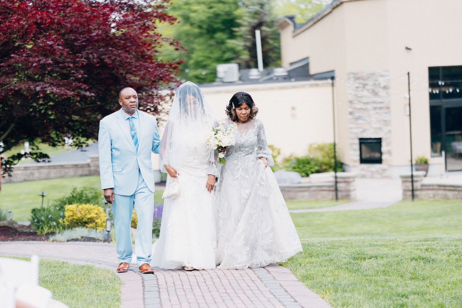 A bride in a white gown walks with two elegantly dressed guests along a garden pathway at a wedding.