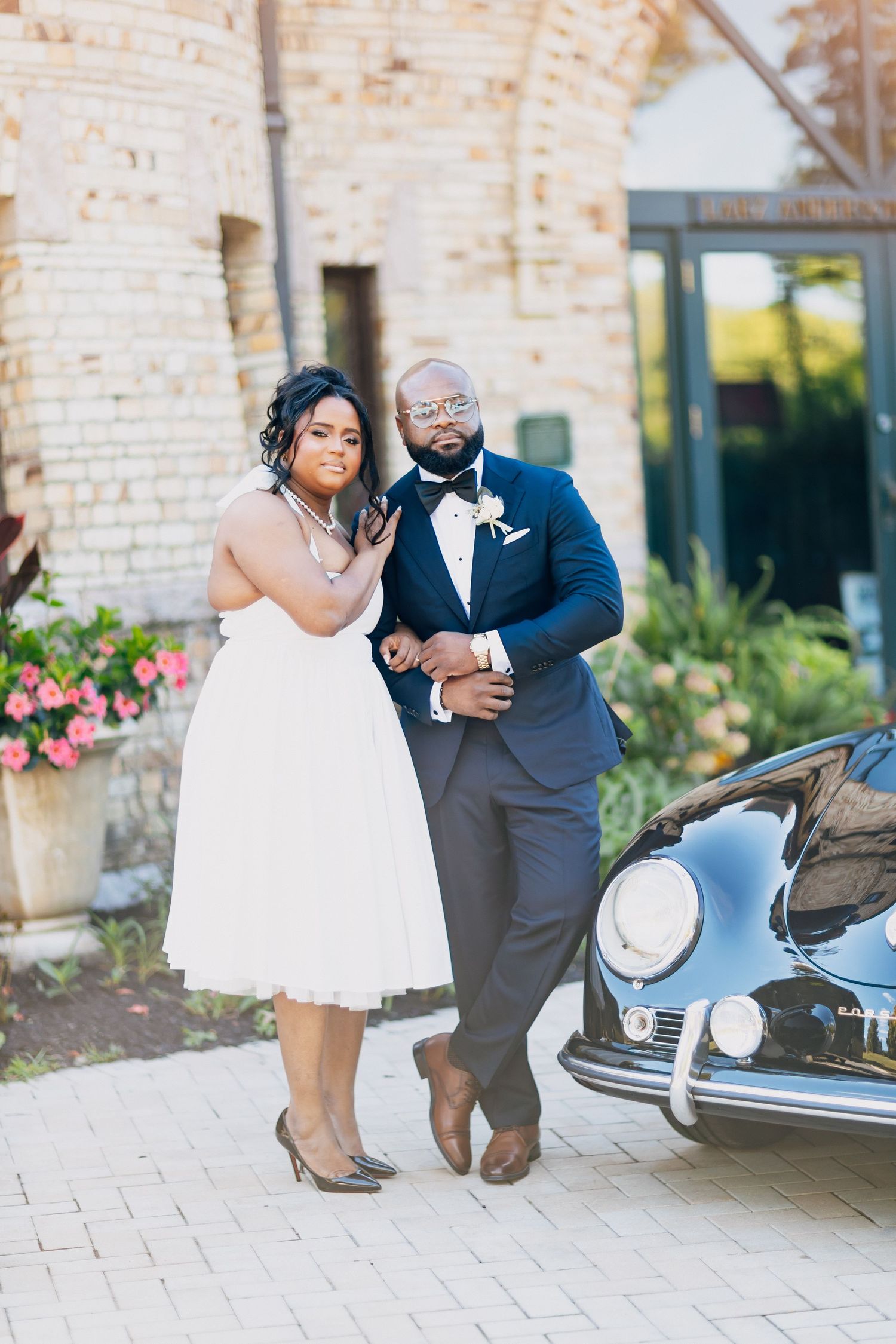 Black couple in wedding attire posing beside a vintage black Porsche outside a brick building.