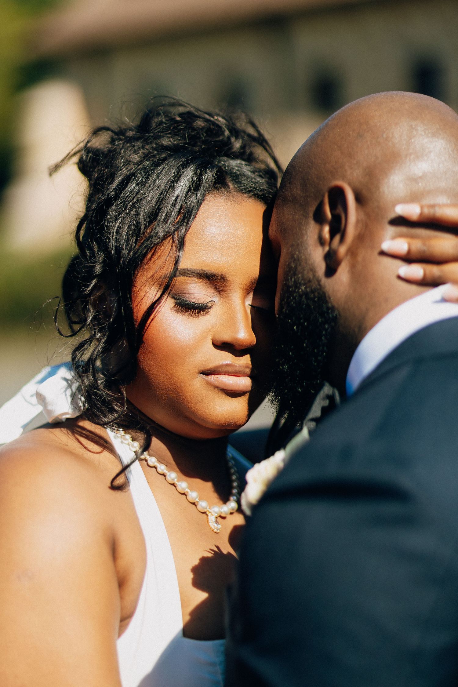 Close-up of a beautiful Black bride with pearl necklace resting her head on her groom's shoulder.