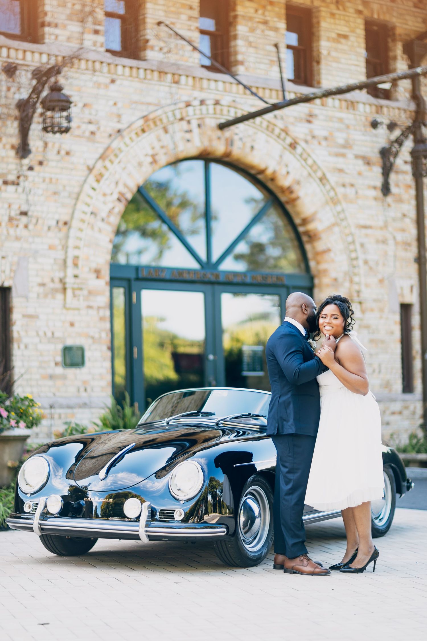 African American couple embraces in front of a classic black Porsche at a brick wedding venue.