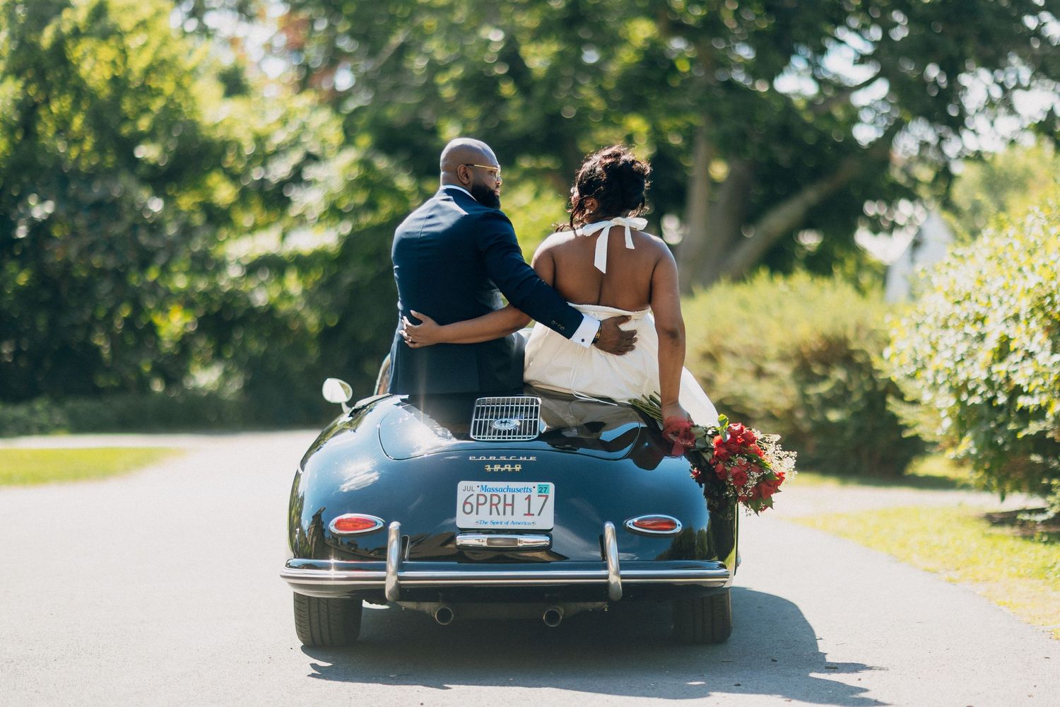 Black bride and groom riding away in a classic navy blue vintage convertible car on their wedding day.