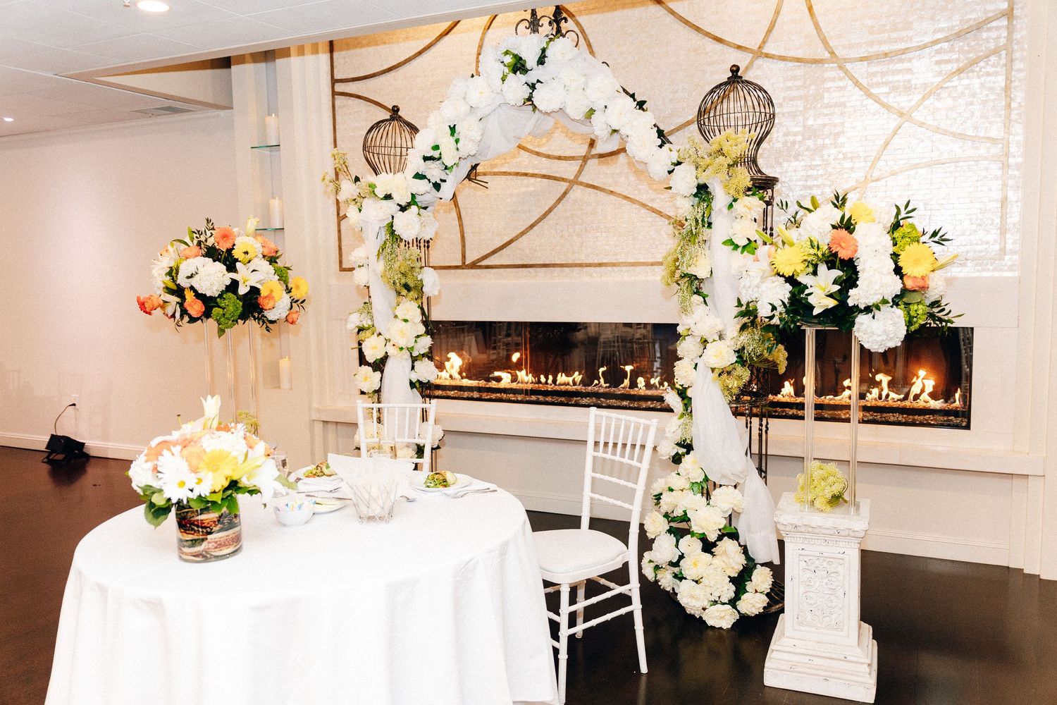 Elegant wedding sweetheart table with floral arch, bird cages, and fireplace backdrop adorned with white and yellow flowers.