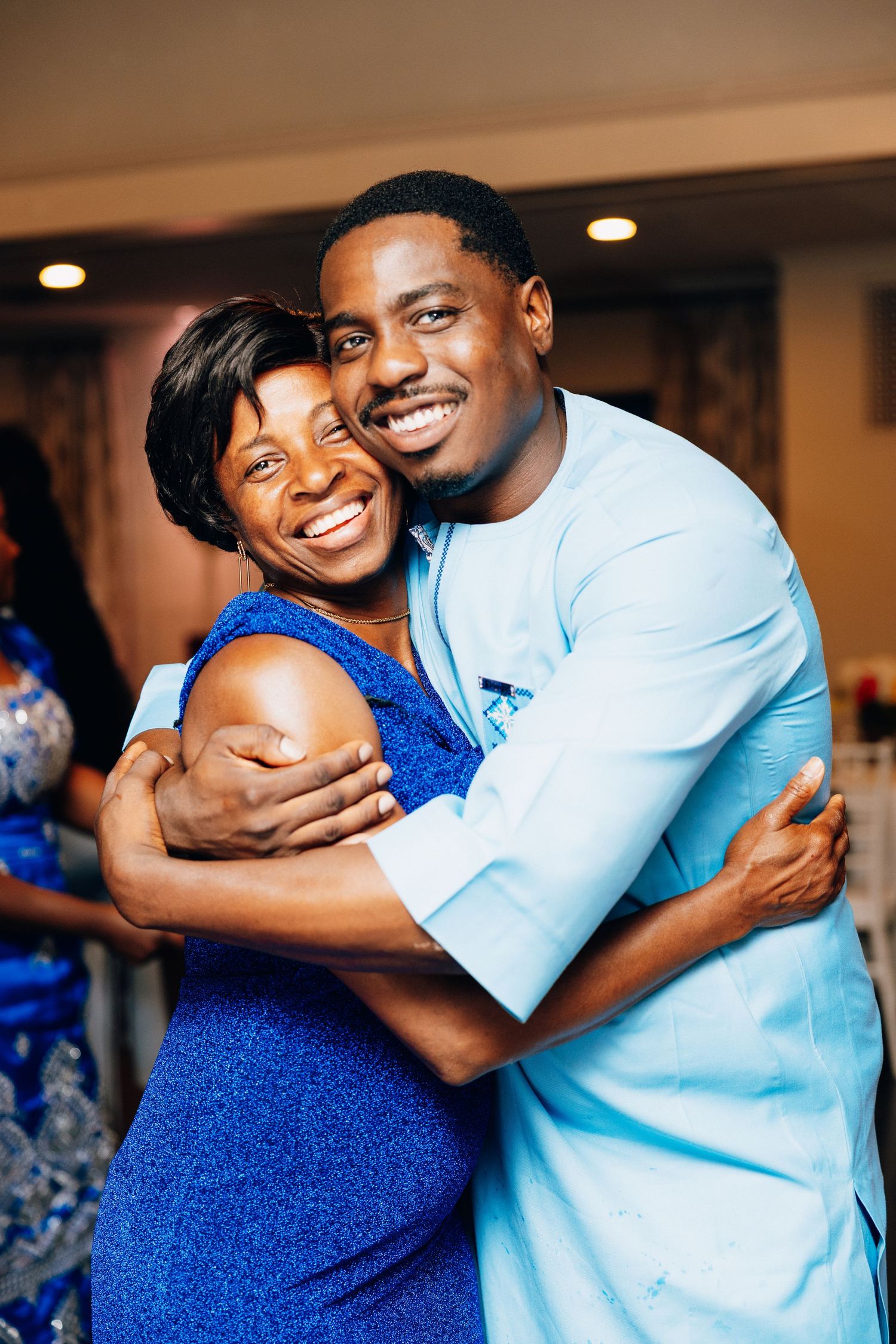 Joyful man in light blue African attire embracing a smiling woman in a blue dress at a celebration.