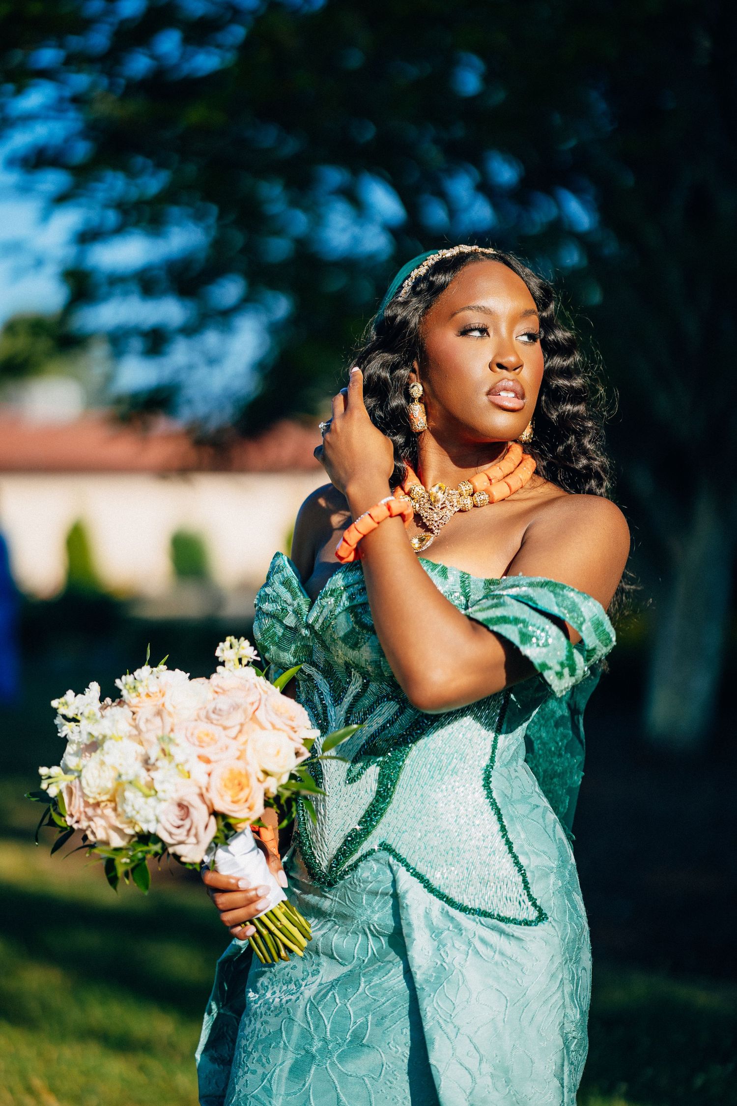 African bride in teal off-shoulder gown holds floral bouquet outdoors.