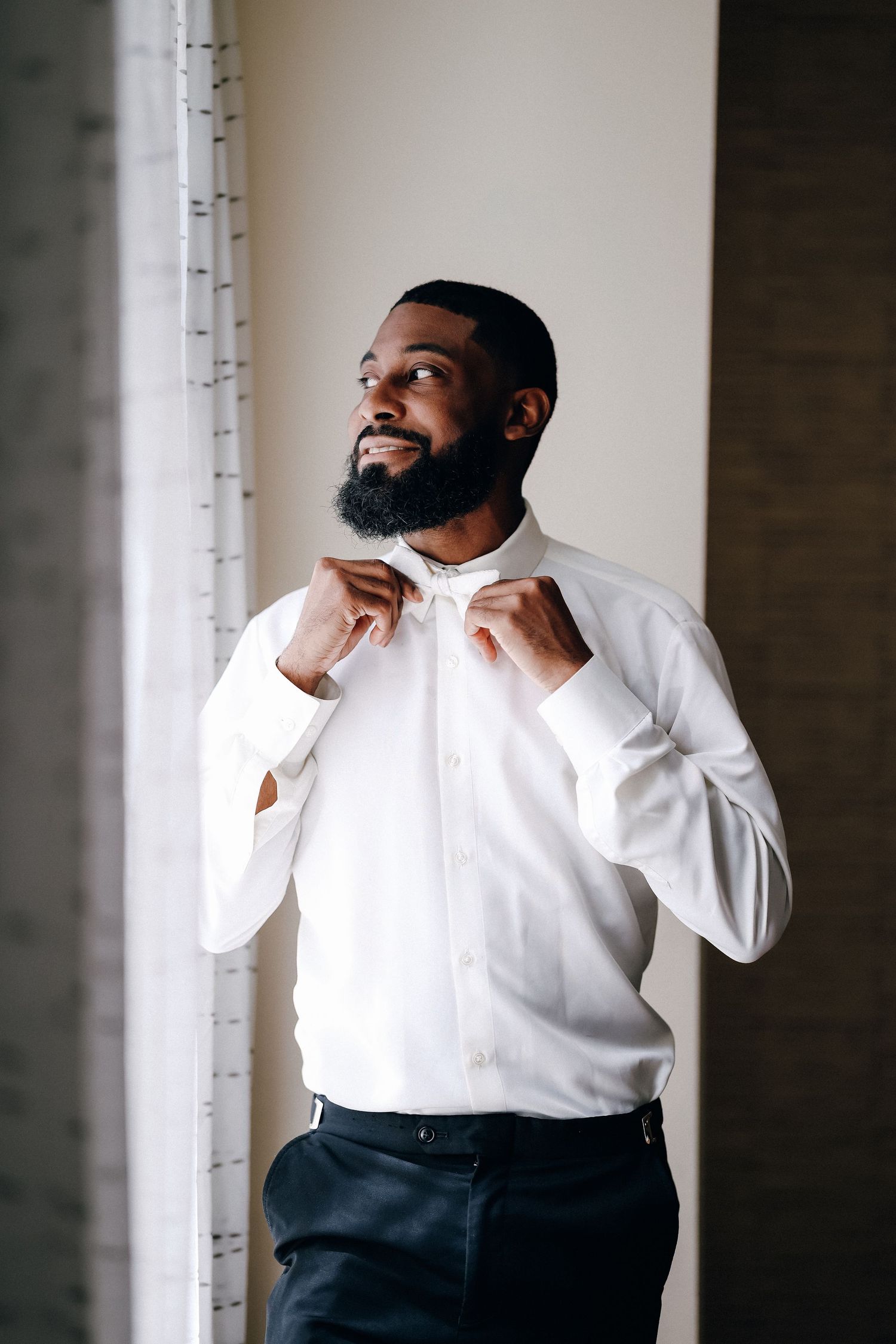 Smiling Black groom adjusting bow tie while wearing white dress shirt before wedding ceremony.