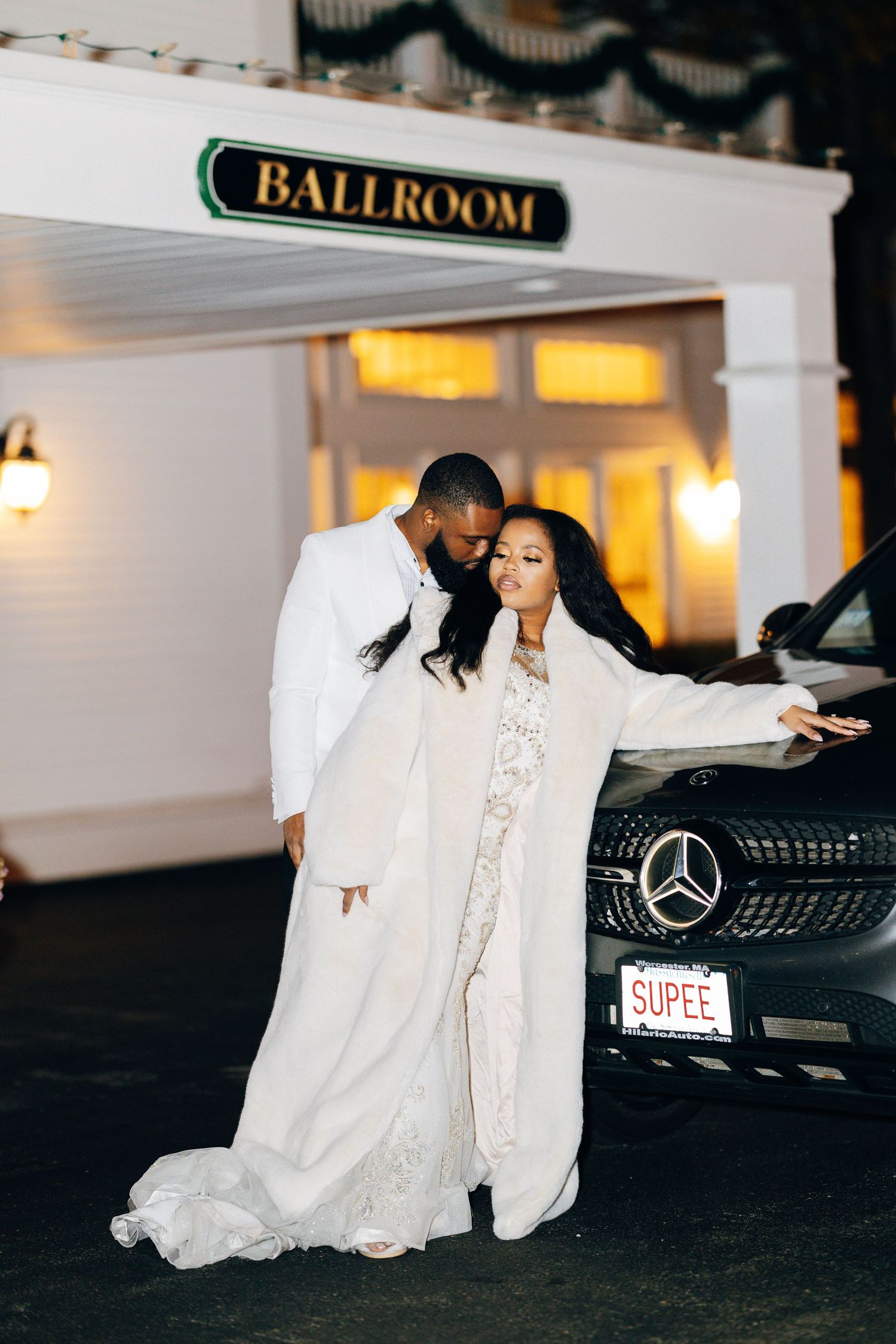Elegant couple in white wedding attire posing by a black Mercedes with SUPEE plate outside a ballroom at night.