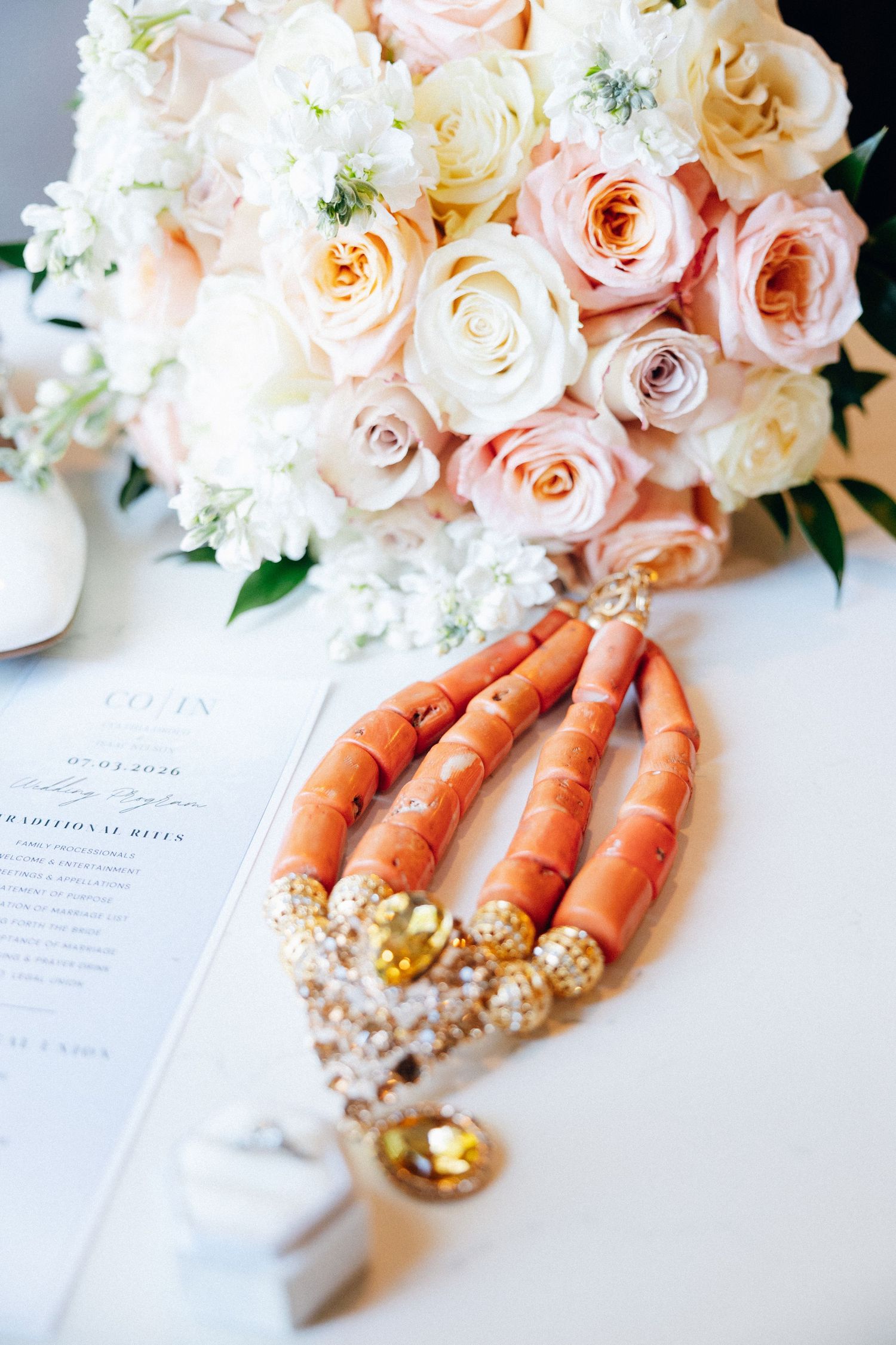 Elegant wedding flat lay with coral beaded necklace, blush pink rose bouquet, and ceremony program on white surface.