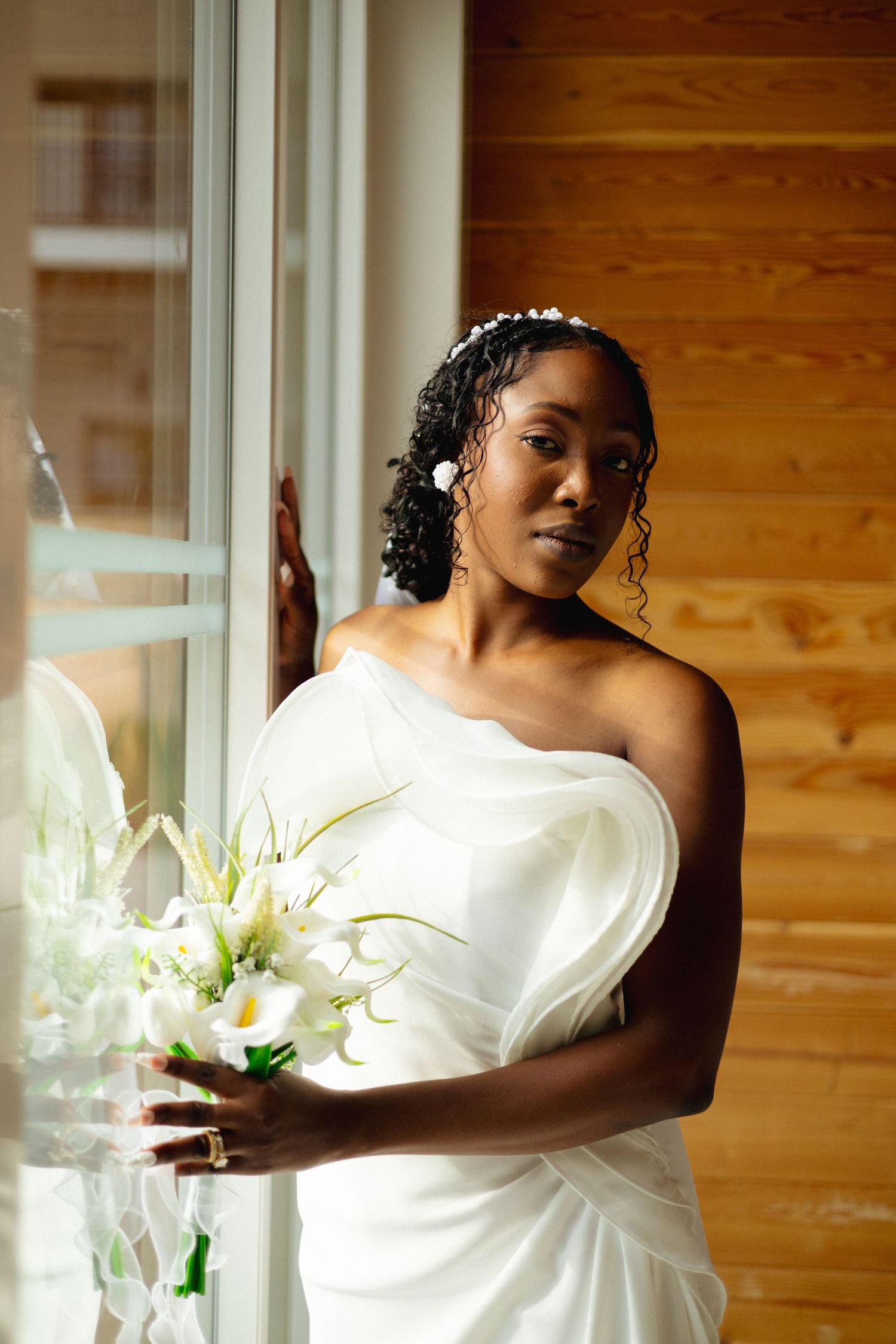 An elegant bride in a white asymmetric gown holds white calla lilies while posing by a large window.