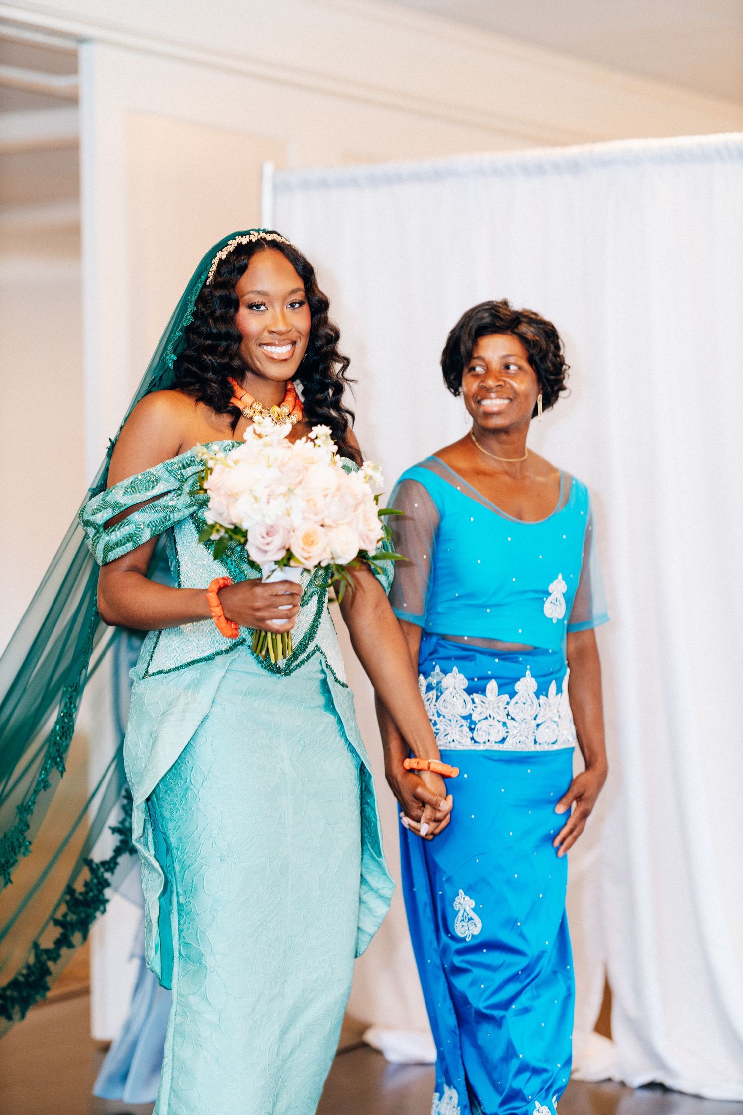 Bride in teal African gown holds bouquet while walking with woman in blue traditional attire.