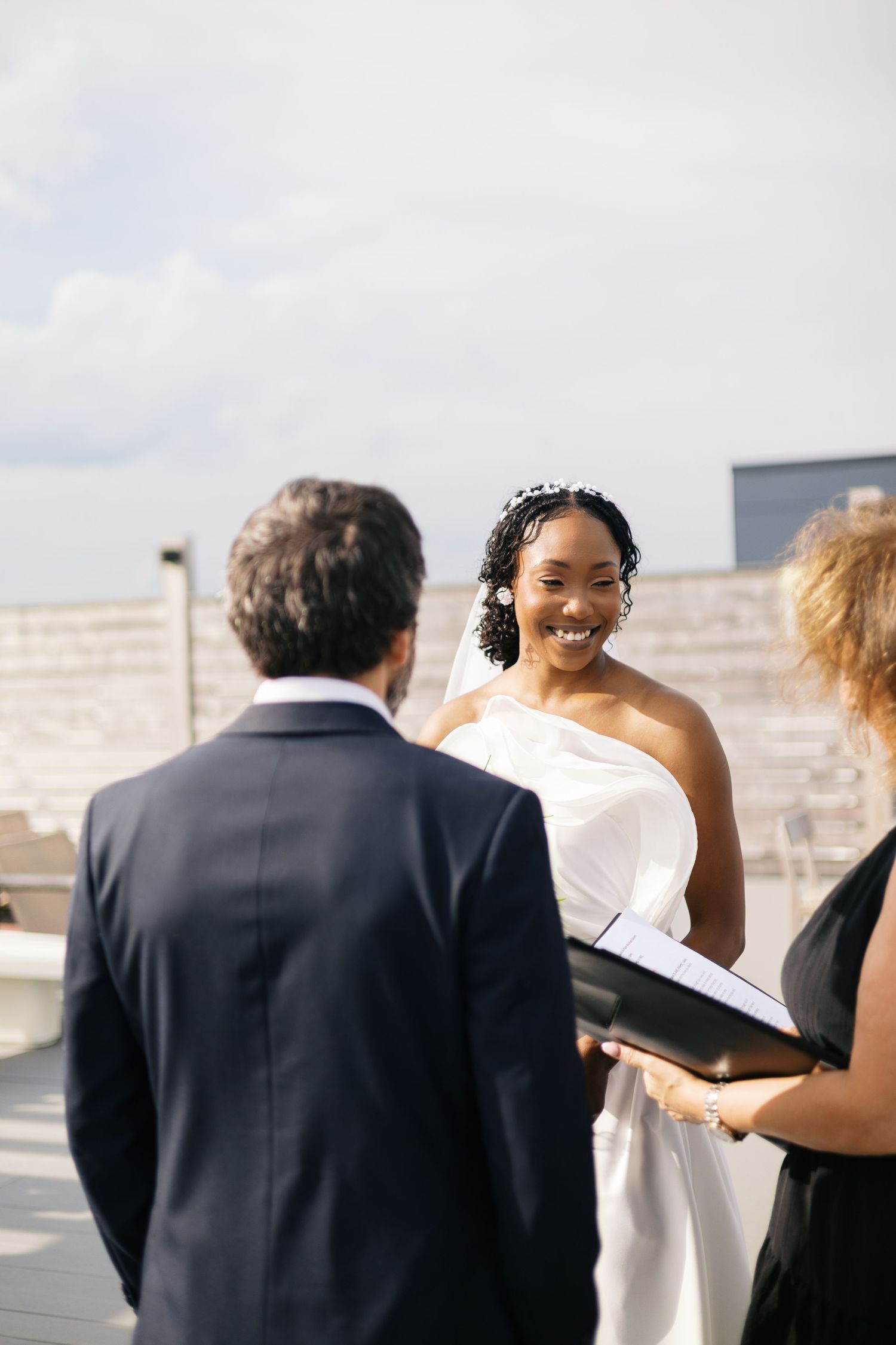 Smiling bride in white dress exchanges vows with groom in navy suit during outdoor rooftop wedding ceremony.
