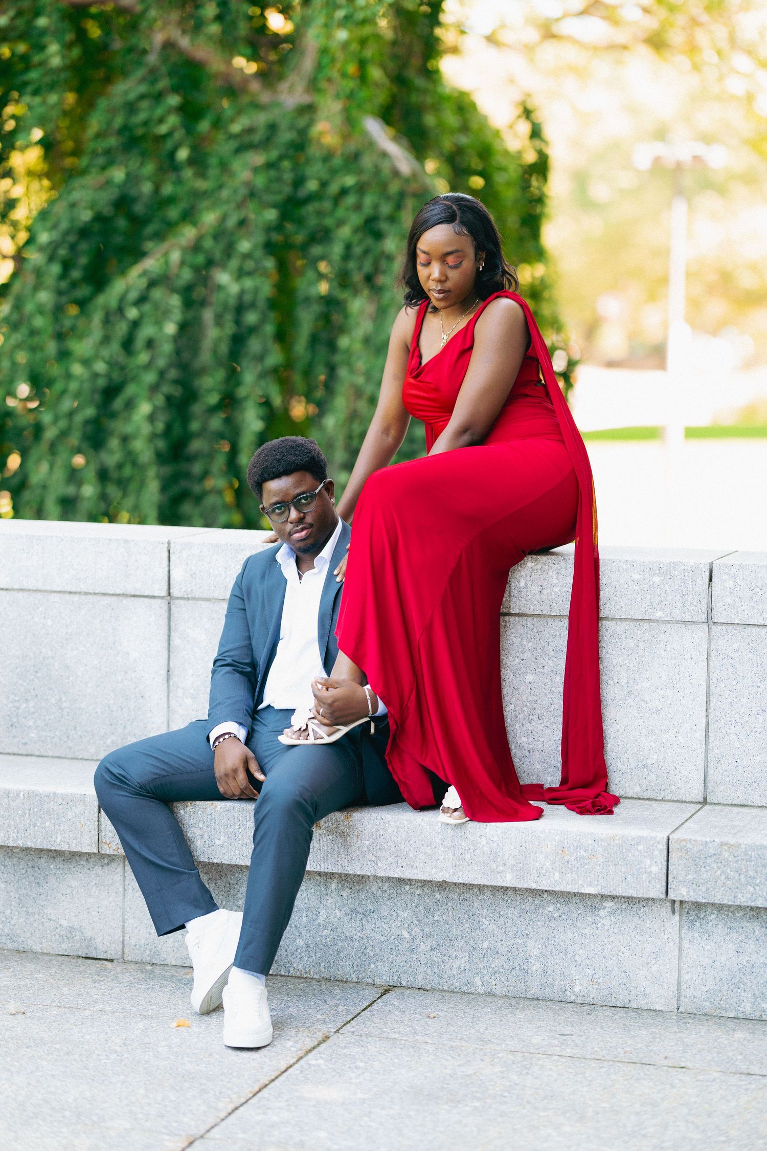 A couple poses on a stone bench outdoors, woman in red gown and man in gray suit with white sneakers.