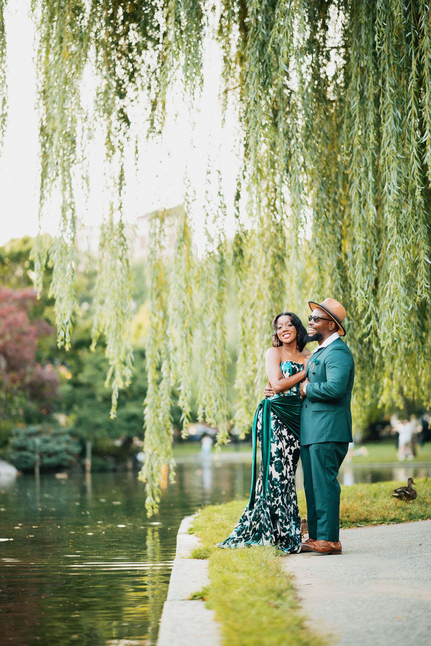 Joyful couple in elegant green attire embracing under weeping willow tree by serene pond.