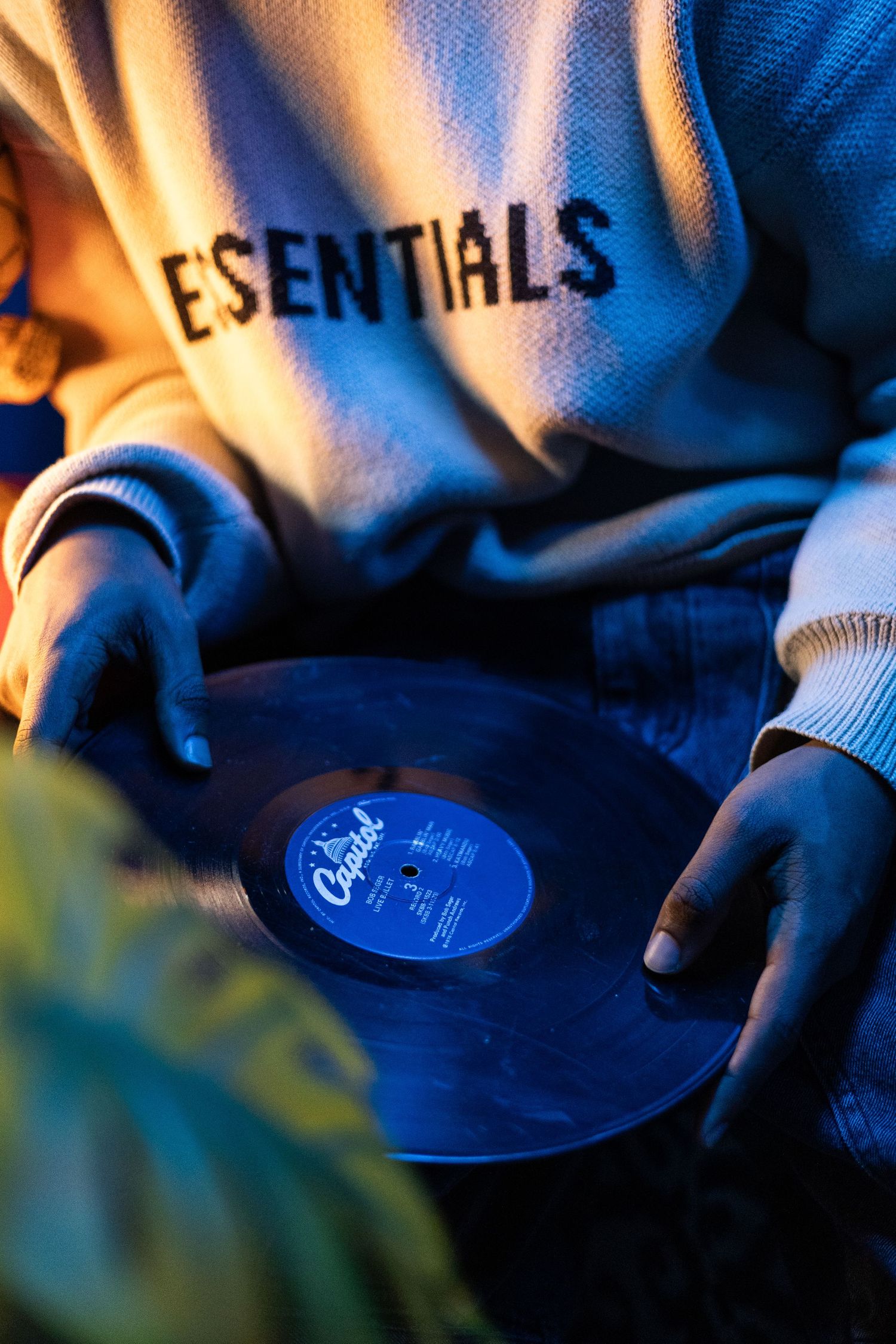 Person in Essentials sweatshirt holding a Capitol Records vinyl record under blue ambient lighting.
