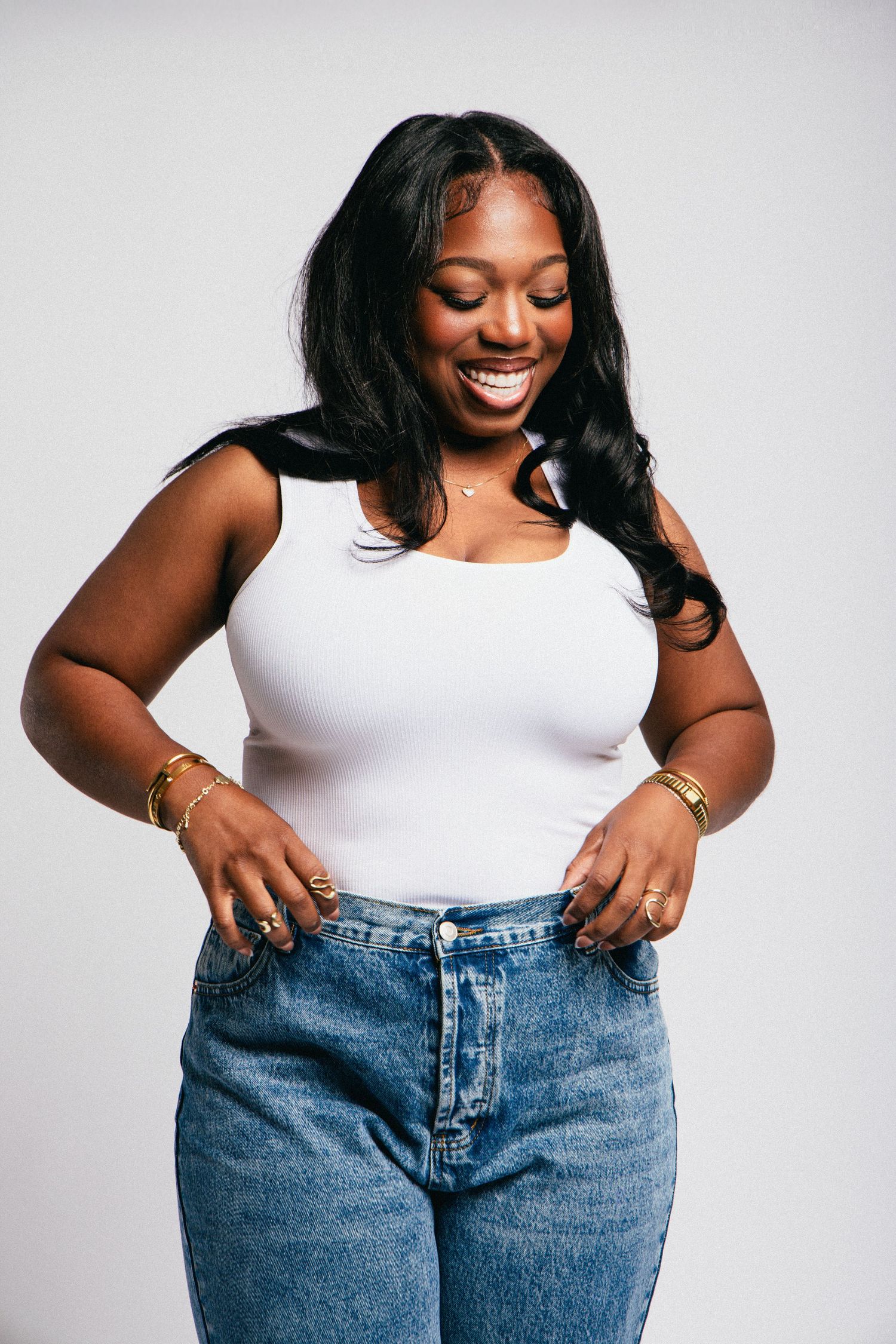 Smiling young Black woman in white tank top and blue jeans, posing confidently with gold jewelry on gray background.