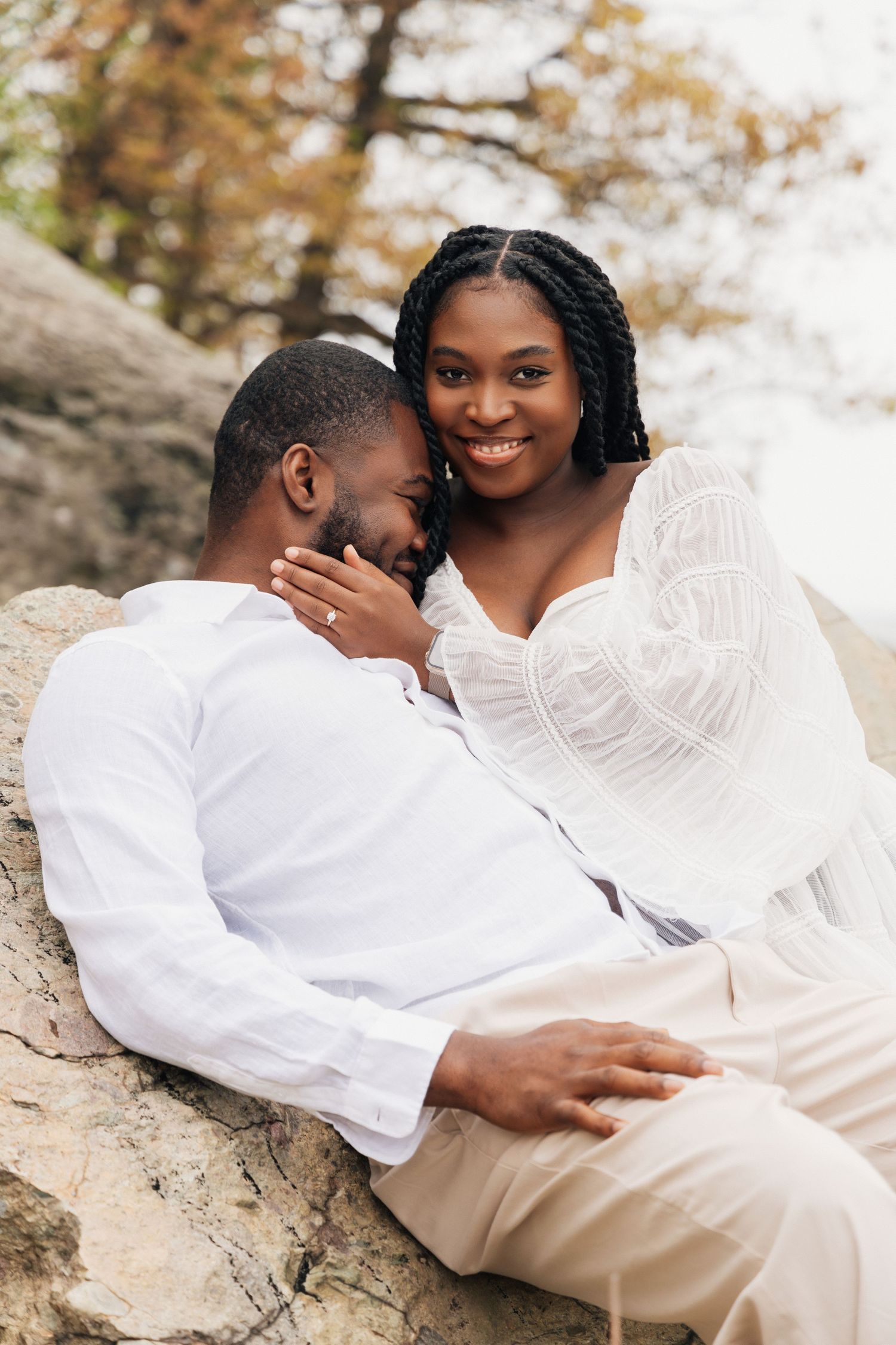 Happy Black couple in white outfits sitting on rocks, smiling woman with braids leaning against mans face outdoors.