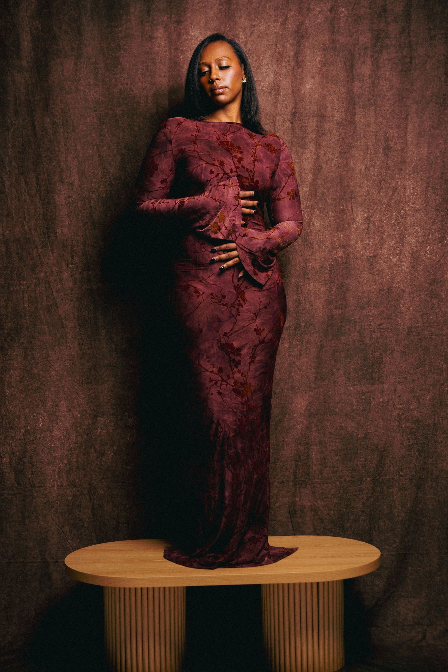 Woman in burgundy floral lace long sleeve gown posing on wooden platform against dark brown backdrop.